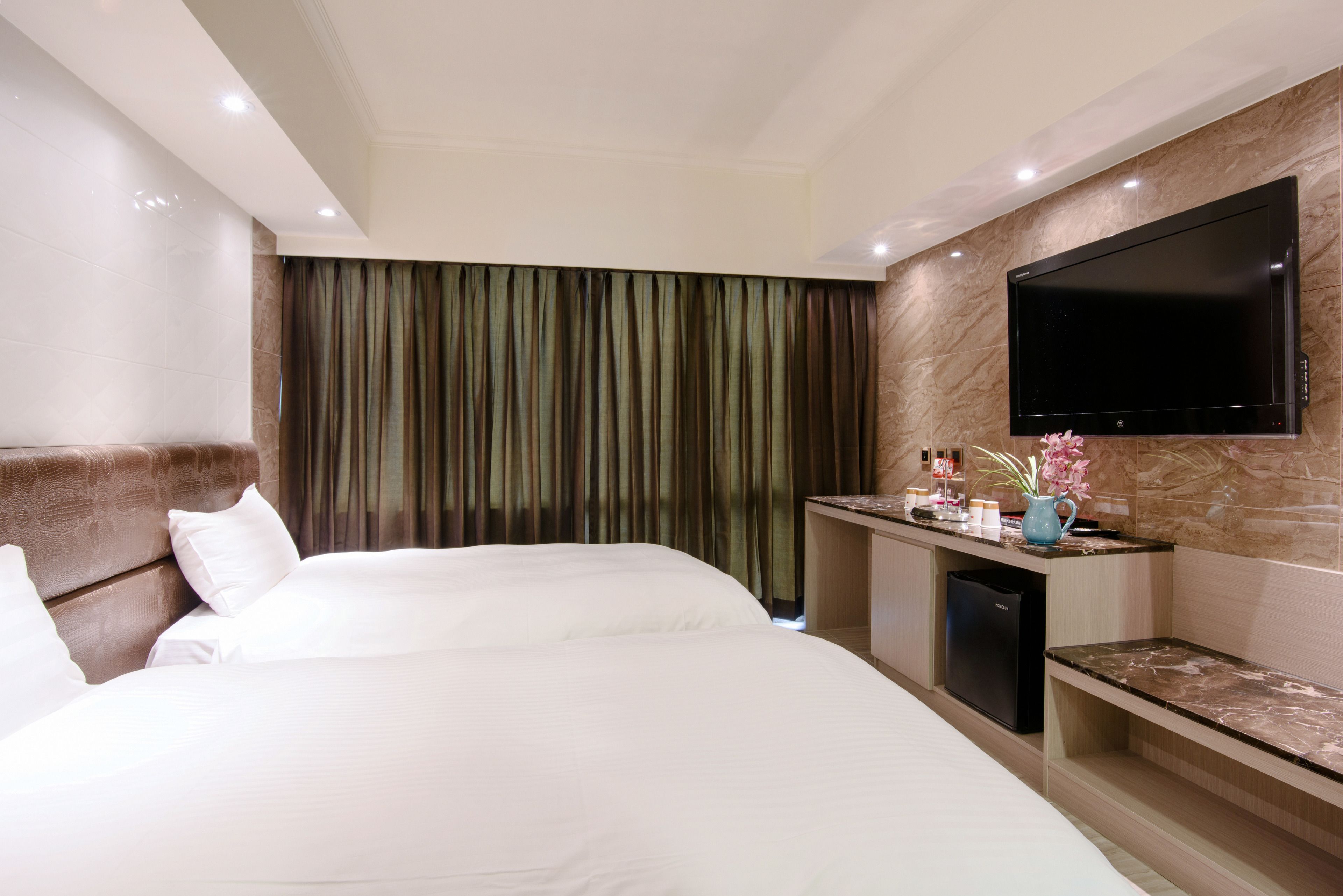 A hotel room with two beds, a flat-screen TV mounted on the wall, a small kitchenette, and a large window with curtains.