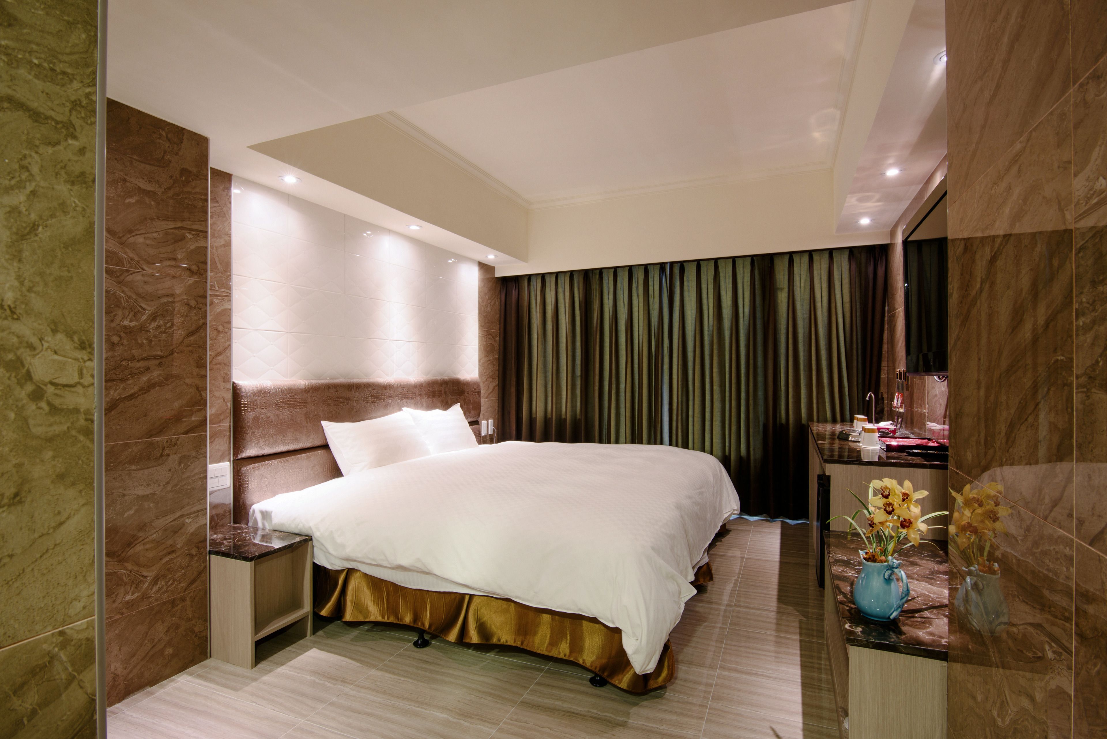 A hotel room with a large bed, a nightstand, a vanity with a mirror, and a vase with flowers.