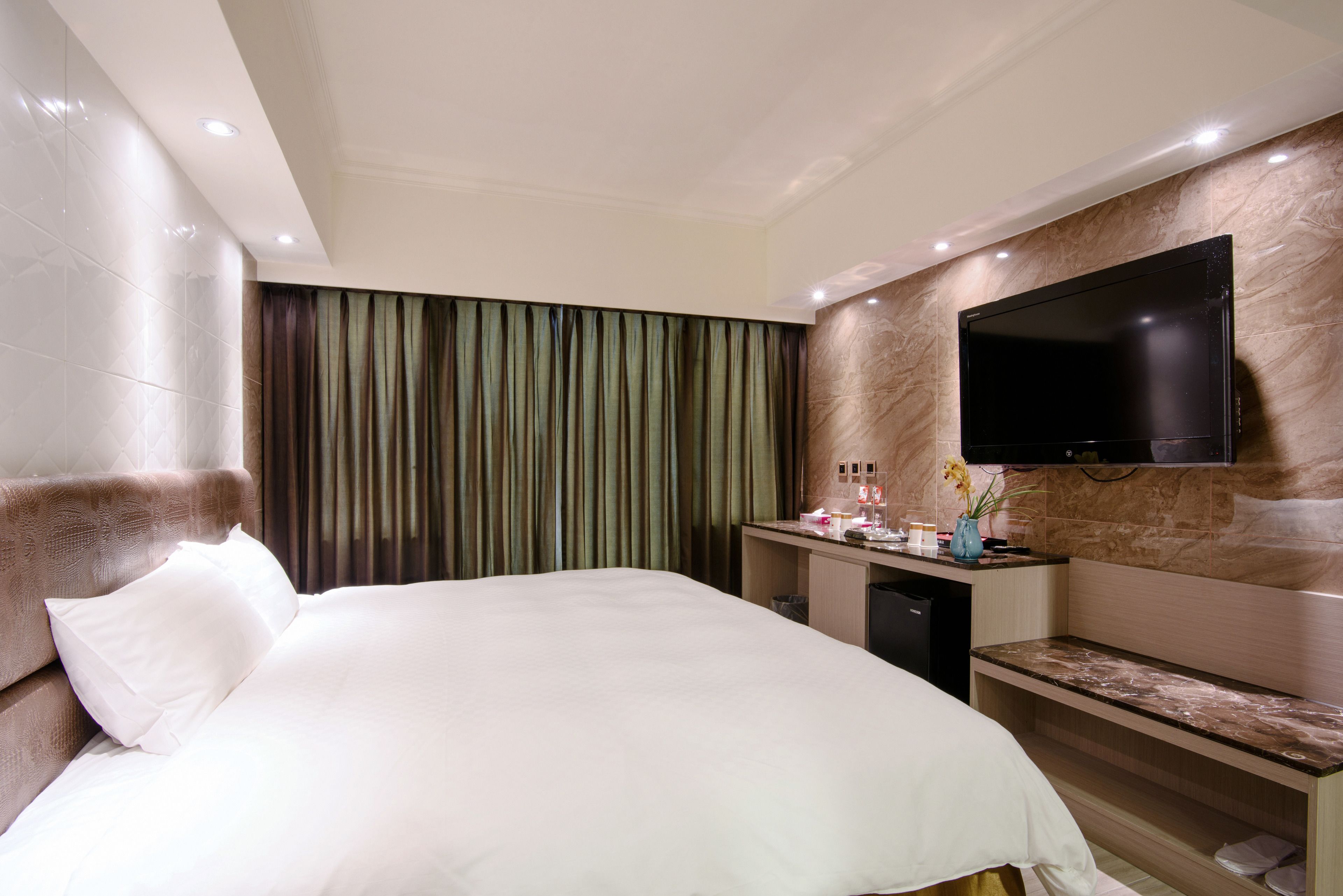 A hotel room with a large bed, a flat-screen TV mounted on the wall, a vanity with a mirror, and a small desk.