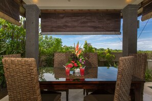 A glass-top table with wicker chairs, a floral centerpiece, and a view of greenery and buildings. - Villa Lidwina by Nagisa Bali (Ungasan)