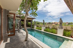 A swimming pool with a clear blue water, surrounded by a stone wall with statues, and a building with a glass door and a covered area. - Villa Lidwina by Nagisa Bali (Ungasan)