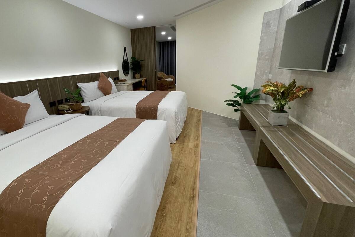 Une chambre d’hôtel avec deux lits, un banc en bois, une télévision et une plante.