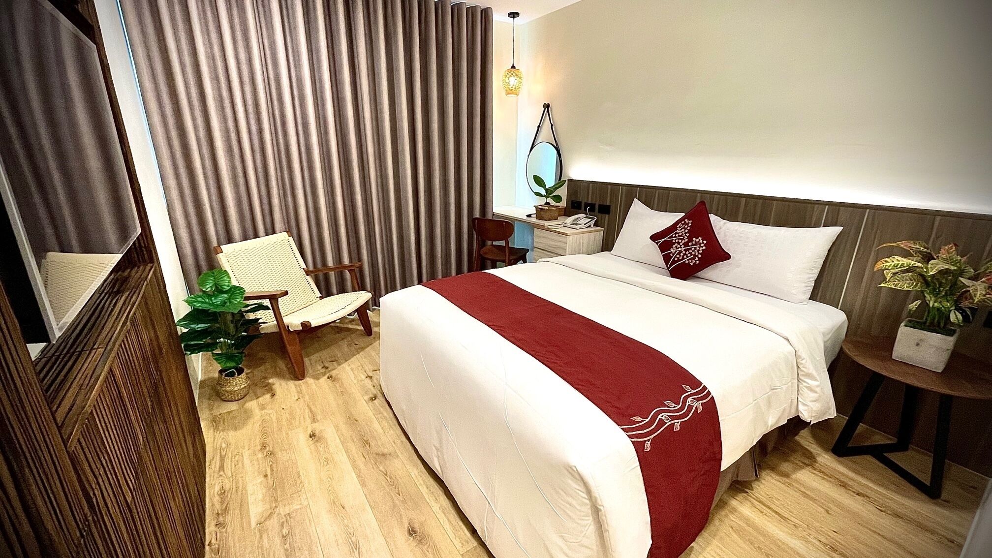 Habitación de hotel con una cama grande, una silla, una mesita de noche con lámpara, una planta en maceta y una ventana con cortinas.