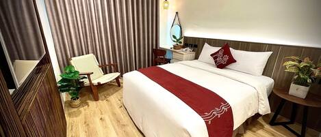 Habitación de hotel con una cama grande, una silla, una mesita de noche con lámpara, una planta en maceta y una ventana con cortinas.