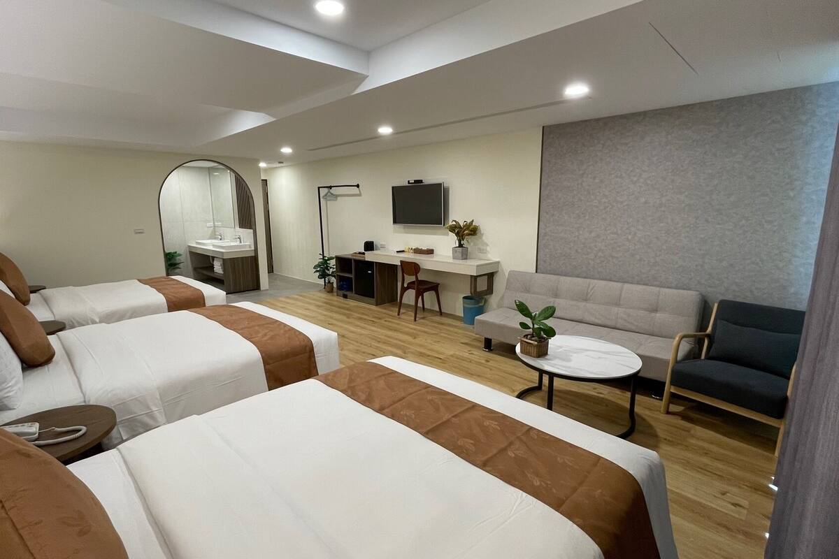 Une chambre d’hôtel avec deux lits, un canapé, une petite table, un bureau, une télévision et une salle de bain avec un miroir et un lavabo.
