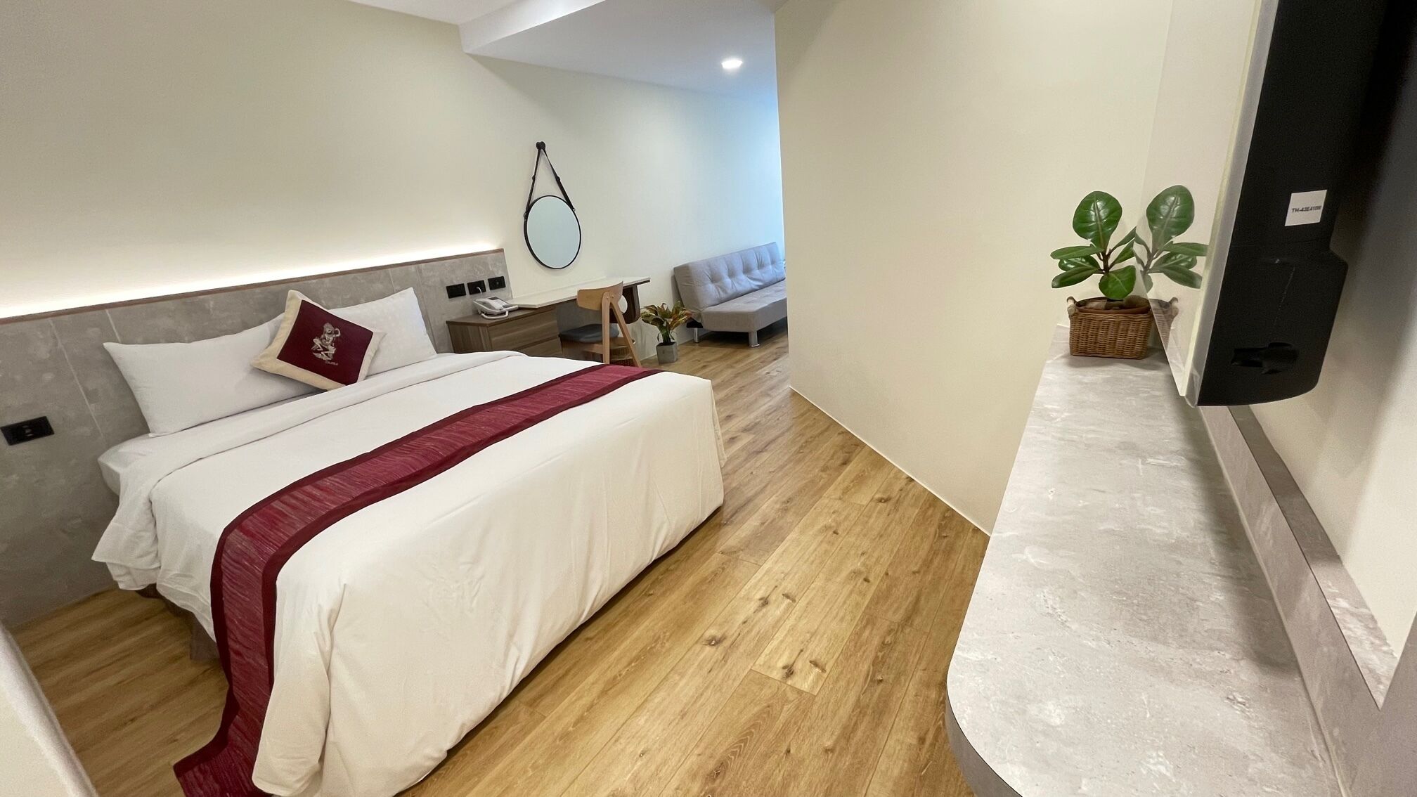 A modern hotel room with a large bed, a bedside table, a nightstand, a chair, a small table with a plant, and a wall-mounted TV.