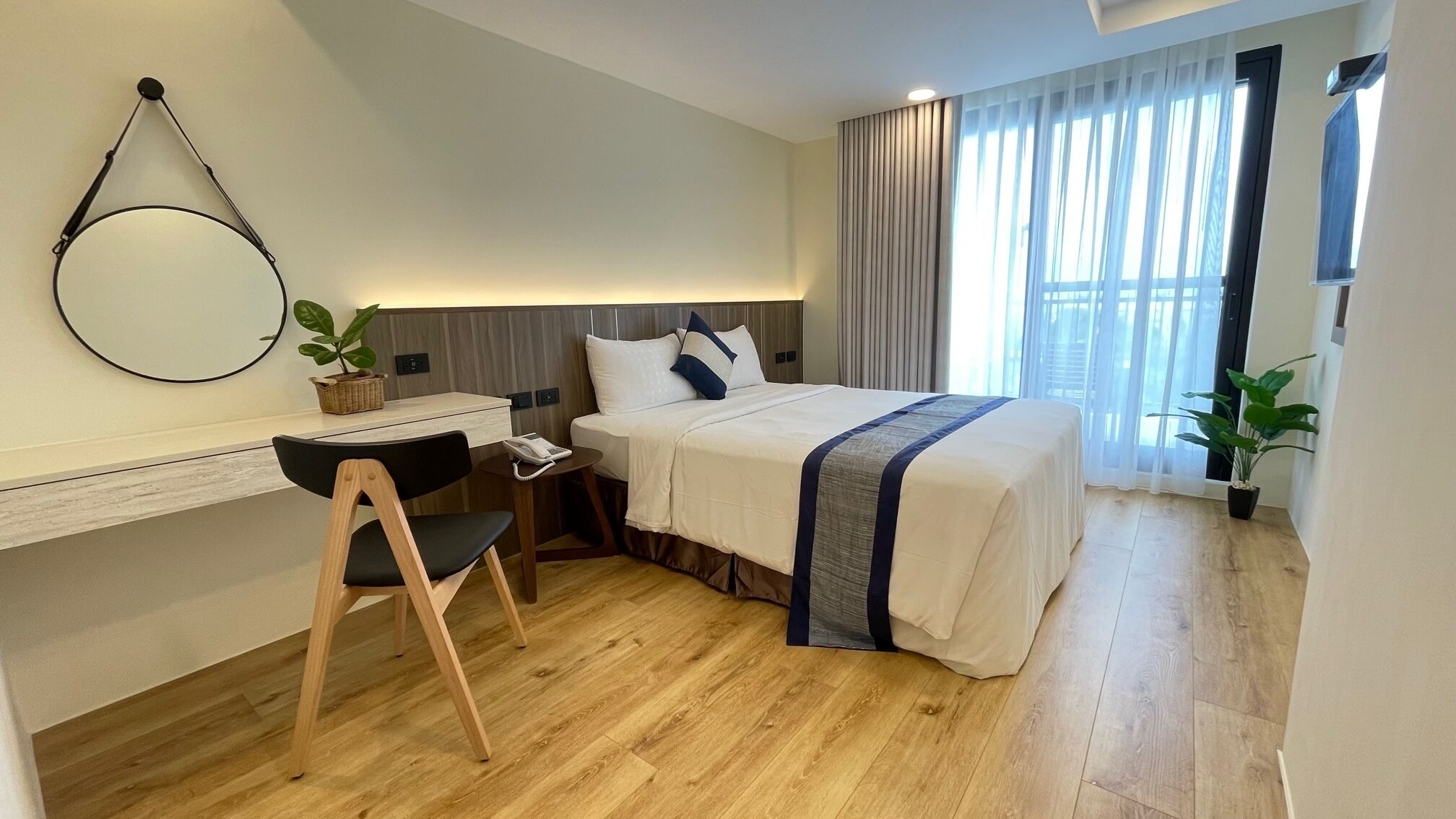 A modern hotel room with a wooden floor, a bed with a blue and white bedspread, a desk with a chair, a phone, and a potted plant.