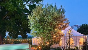 Une piscine avec une terrasse carrelée, entourée d’arbres et agrémentée d’un espace abrité décoré de guirlandes lumineuses.