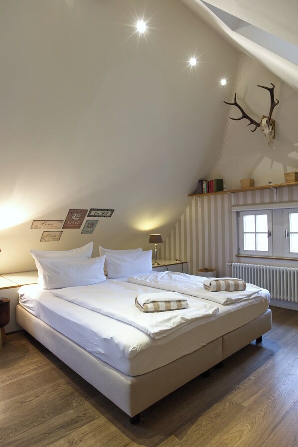 A bedroom with a large bed, a wooden floor, a sloped ceiling, a mounted deer head, and a window with a view.