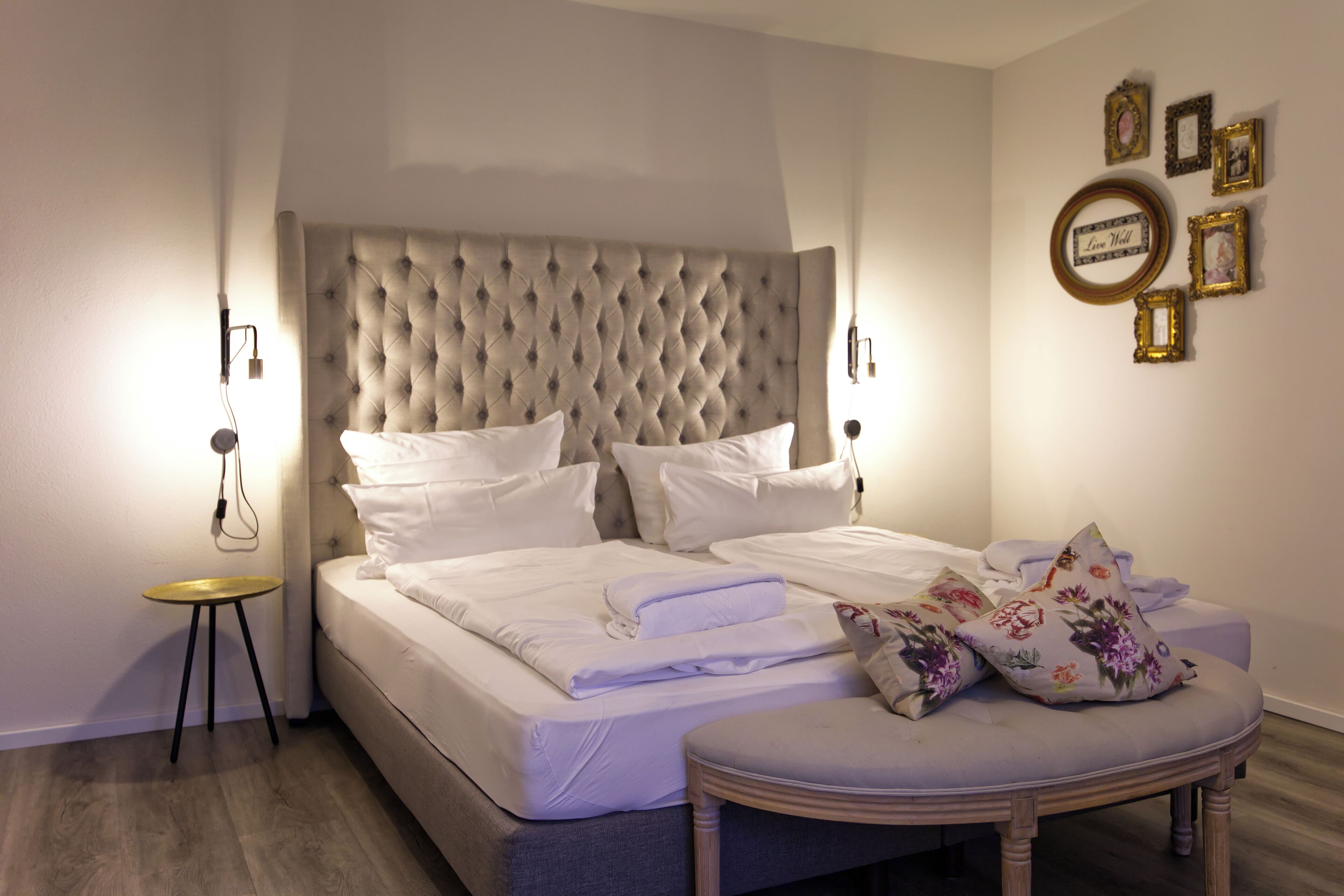 A neatly made bed with white linens and a tufted headboard, a small round wooden table, and a bench with floral cushions.