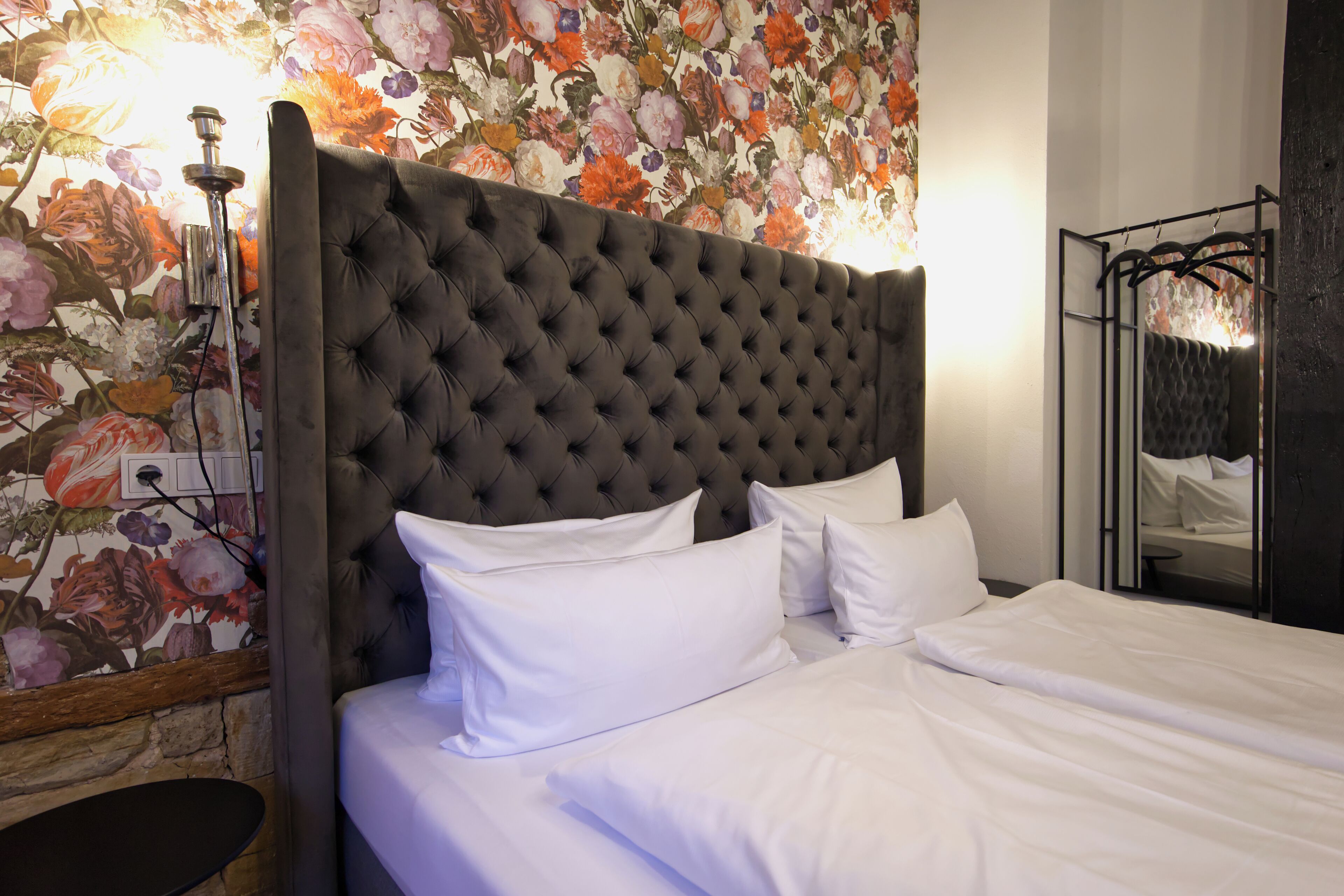 A bed with white linens, a tufted headboard, and a floral wallpaper backdrop.