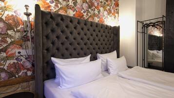 A bed with white linens, a tufted headboard, and a floral wallpaper backdrop.