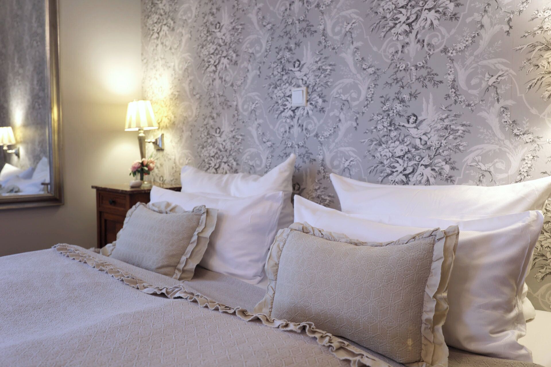 Una cama bien hecha con un cubrelecho fruncido, un estampado floral en el papel tapiz y una mesita de noche con una lámpara y flores.