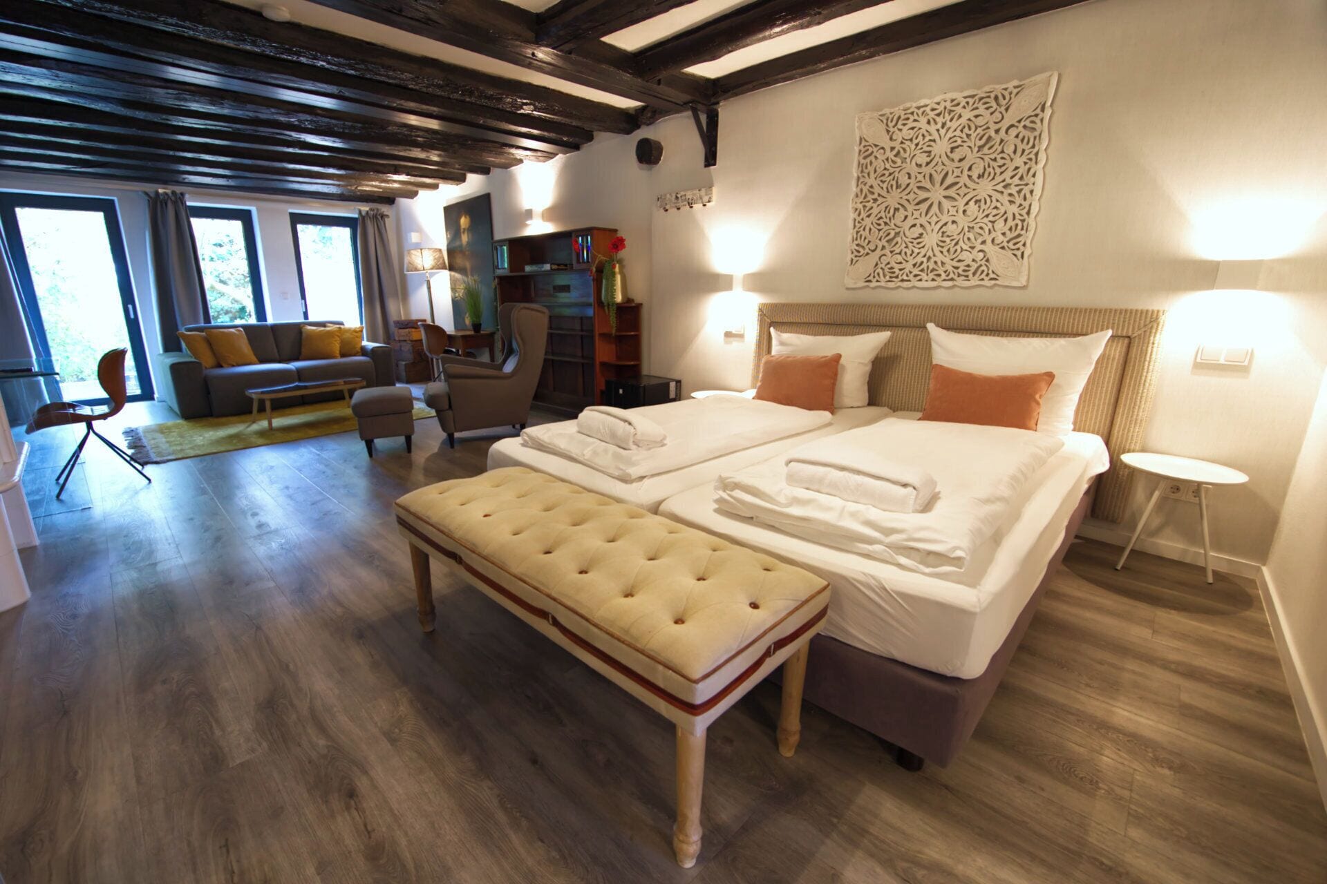 Ein Hotelzimmer mit einem großen Bett, einer Couch, einem Sessel und einem Tisch. Es gibt Fenster und eine Decke mit freiliegenden Balken.