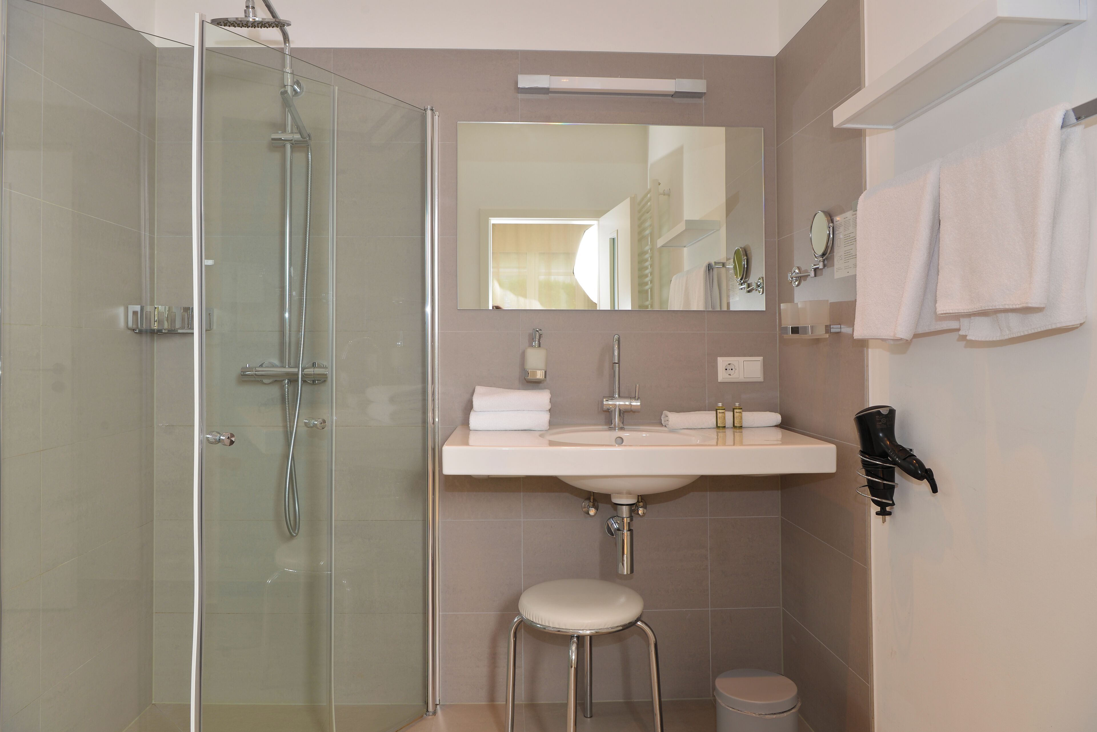 Ein modernes Badezimmer mit einer gläsernen Duschkabine, einem Waschbecken mit Spiegel und einem an der Wand befestigten Föhn.