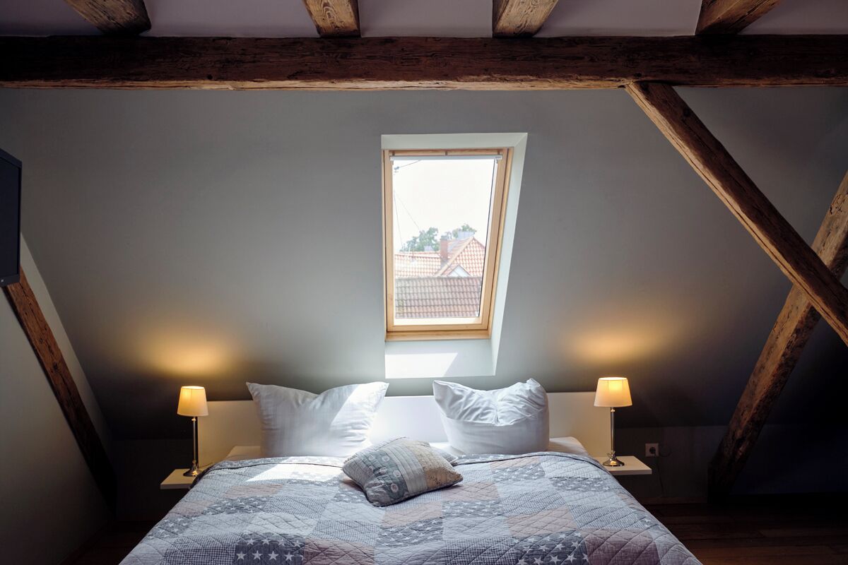 Ein Schlafzimmer mit schrägen Wänden, einem Fenster, zwei Nachttischlampen und einem Bett mit einer Dessin-Quiltdecke und Kissen.