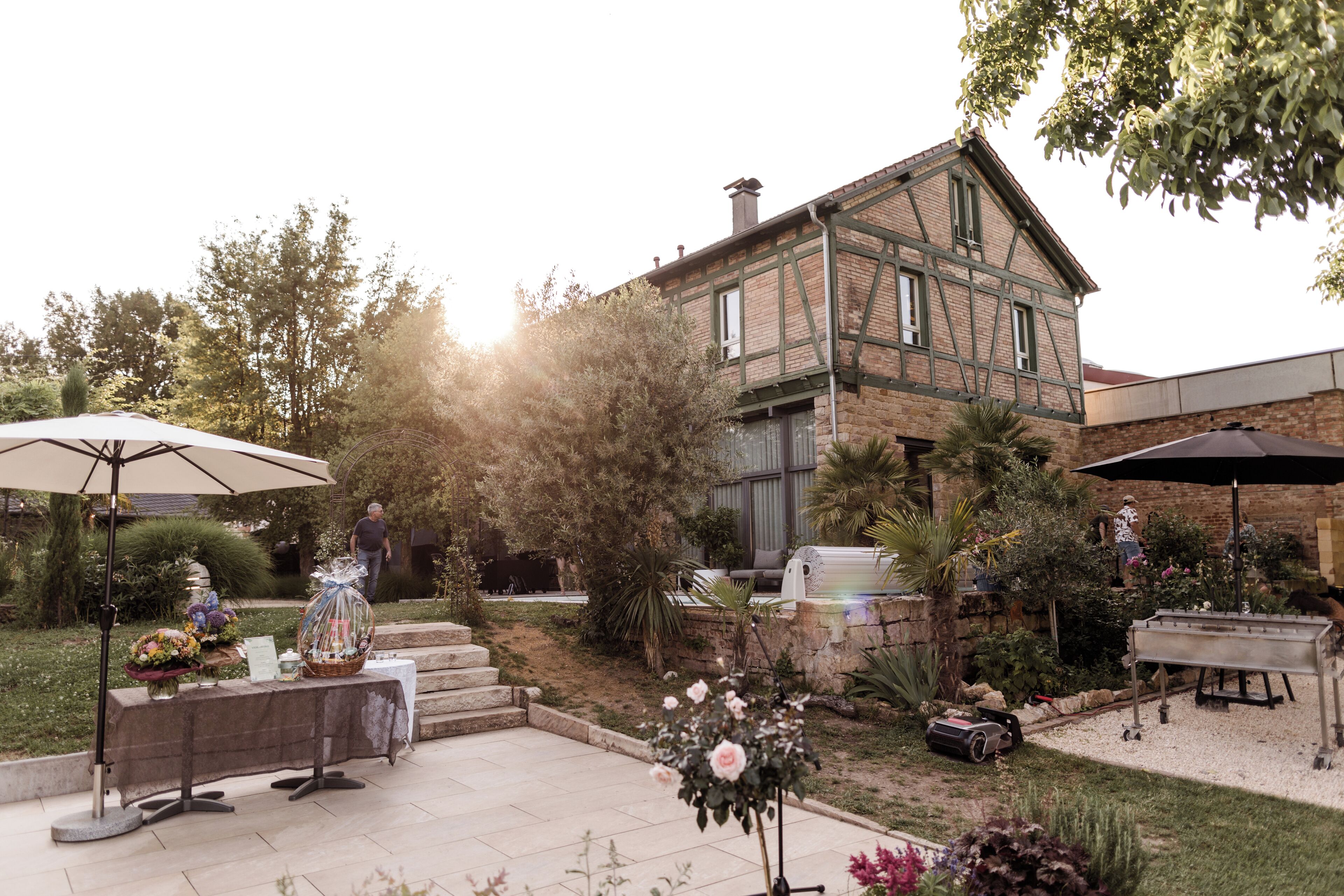 Un espace patio aménagé avec une table, des chaises et des parasols, entouré d’un jardin et d’un bâtiment à la façade en briques.