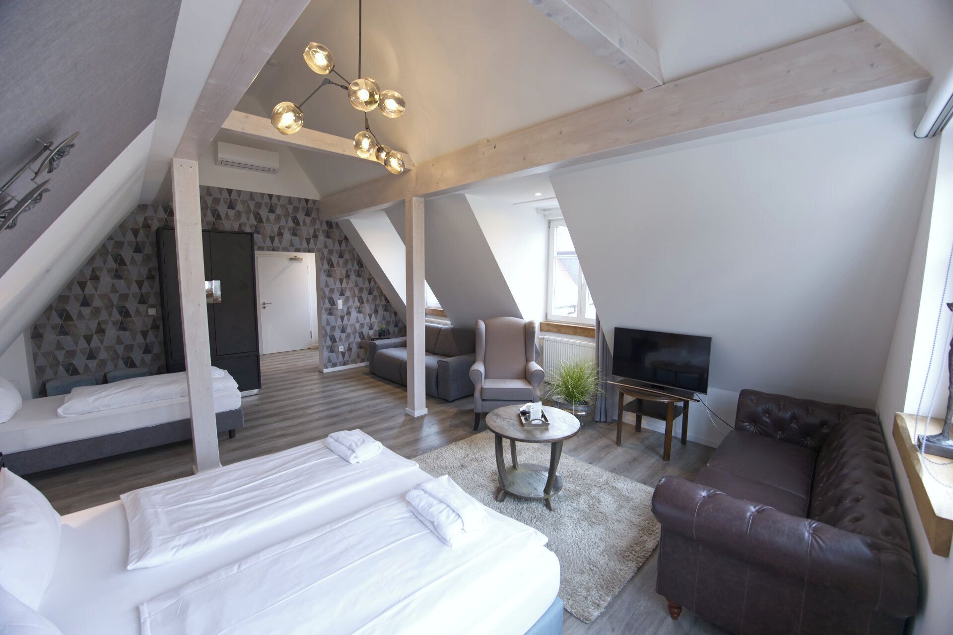 A modern loft bedroom with a large bed, a seating area with a sofa and armchair, a TV, and a dining table.