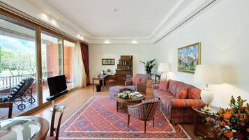A spacious living room with a large sofa, a coffee table, and a television. There is a balcony with outdoor furniture and a mountain view.