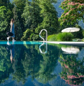 Une piscine aux eaux cristallines, dotée d’un escalier en métal, entourée d’une végétation luxuriante et surmontée d’un parasol blanc.