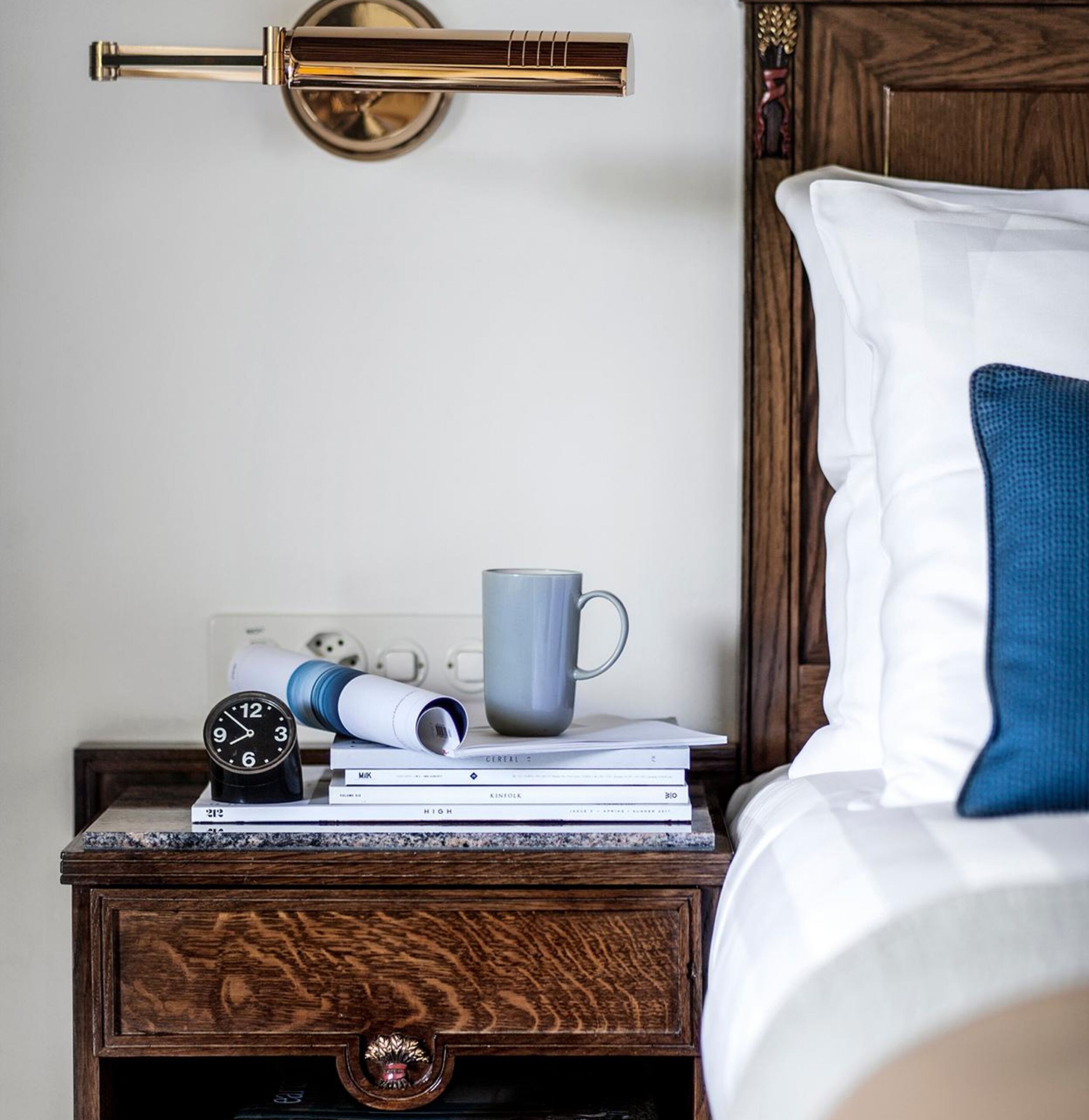 Un letto rifatto con un cuscino blu, un comodino con una sveglia, una tazza e dei libri.