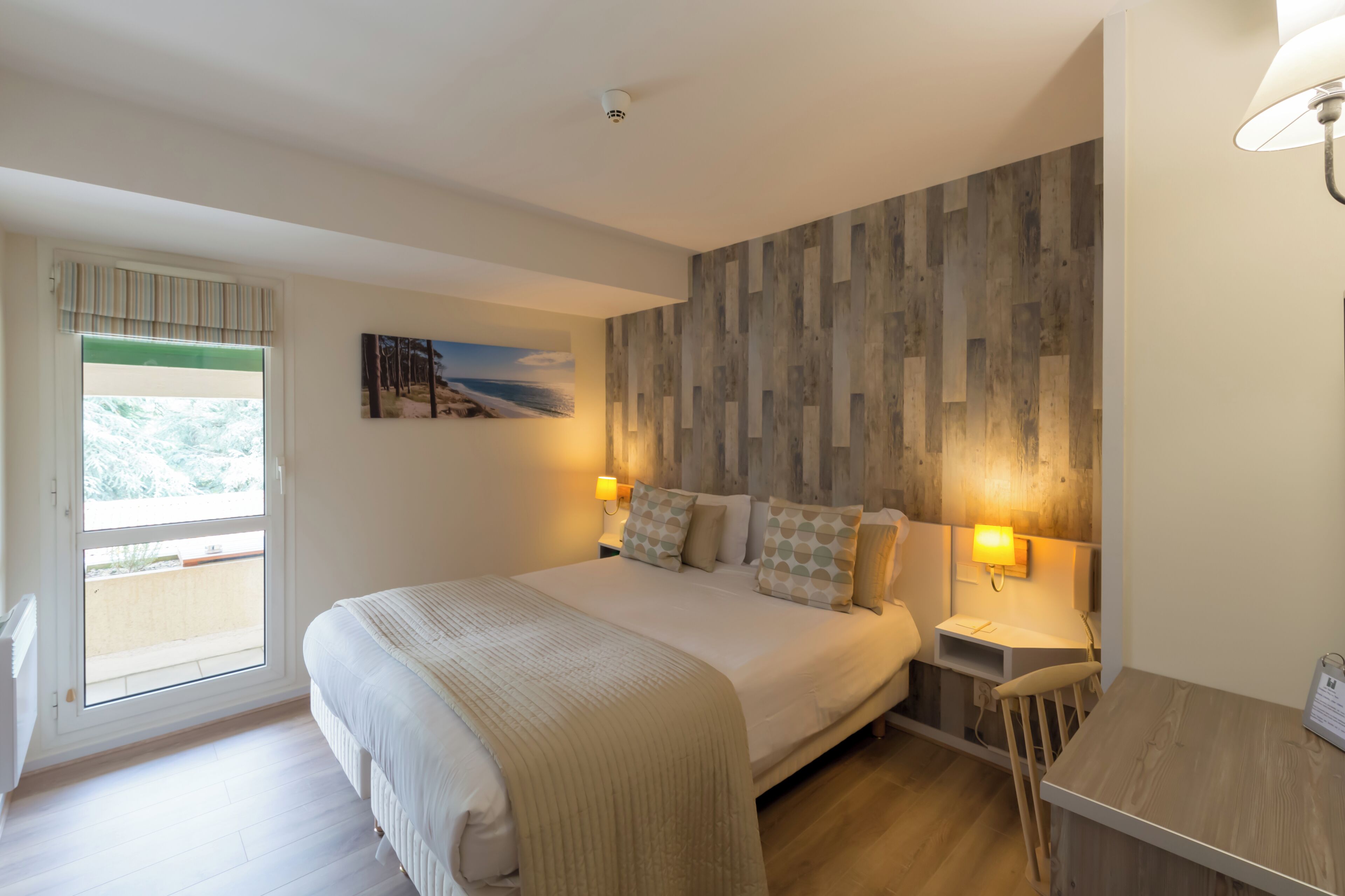 A bedroom with a bed, bedside lamps, a wooden wall panel, a window with a view, and a small table.