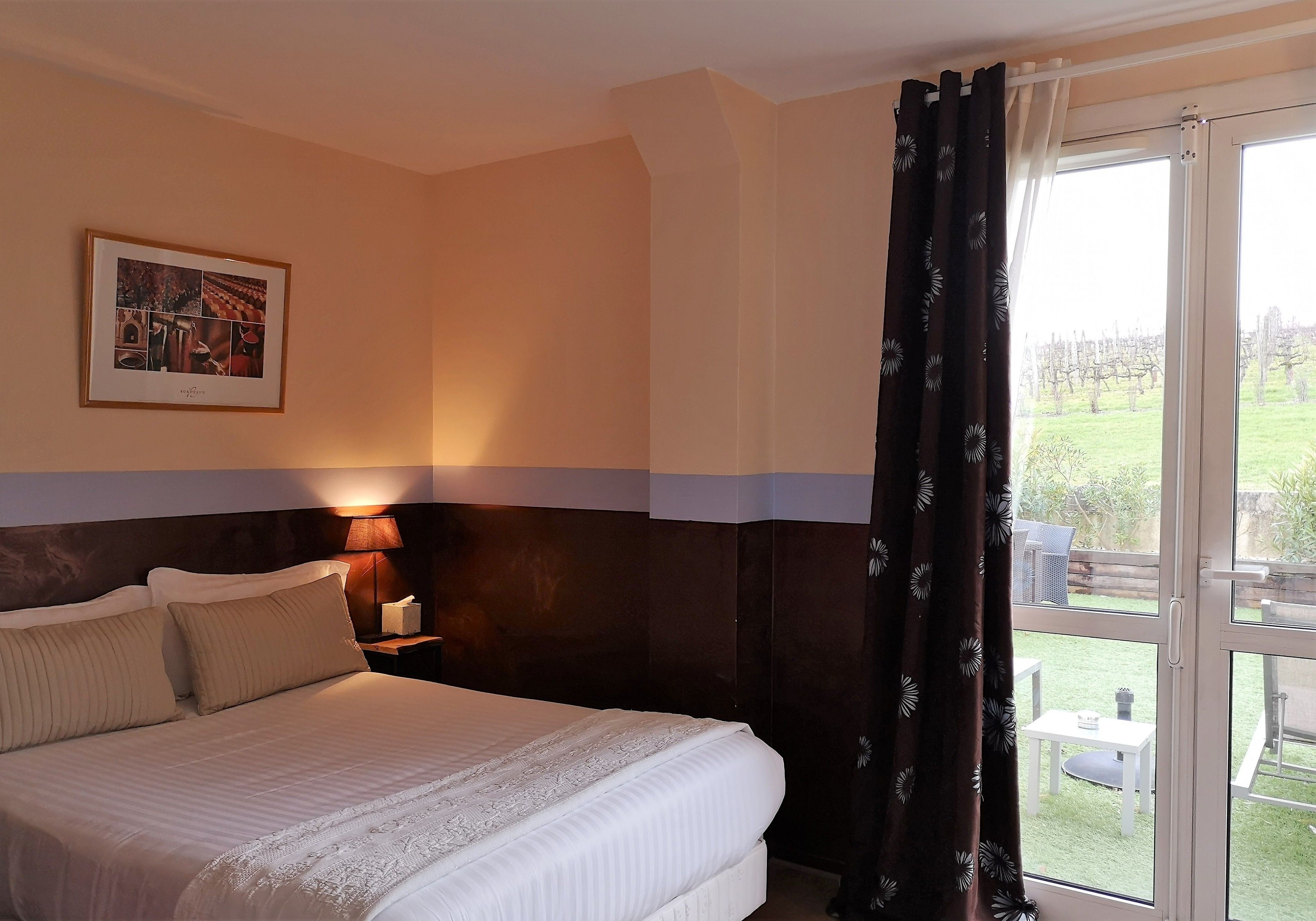 Habitación de hotel con una cama grande, una mesita de noche con lámpara, un cuadro en la pared y vistas al exterior a través de una puerta corredera.