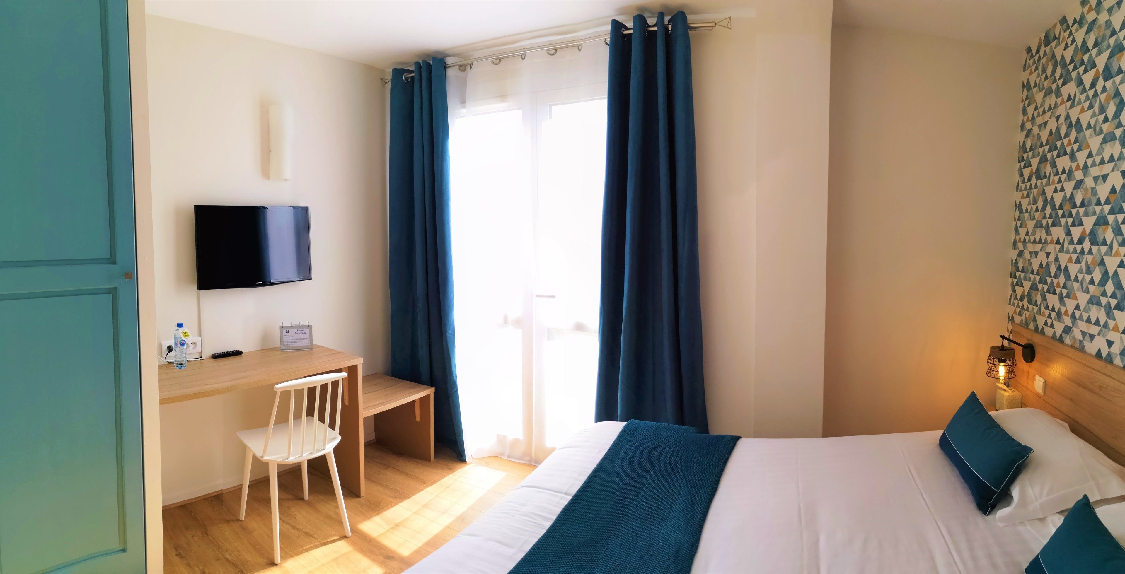 Et hotelværelse med seng, skrivebord, stol, fjernsyn og et vindue med gardiner.