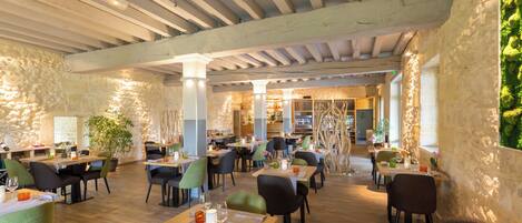 Un restaurante con vigas de madera, paredes de piedra y sillas verdes. Las mesas están preparadas con copas de vino y platos.