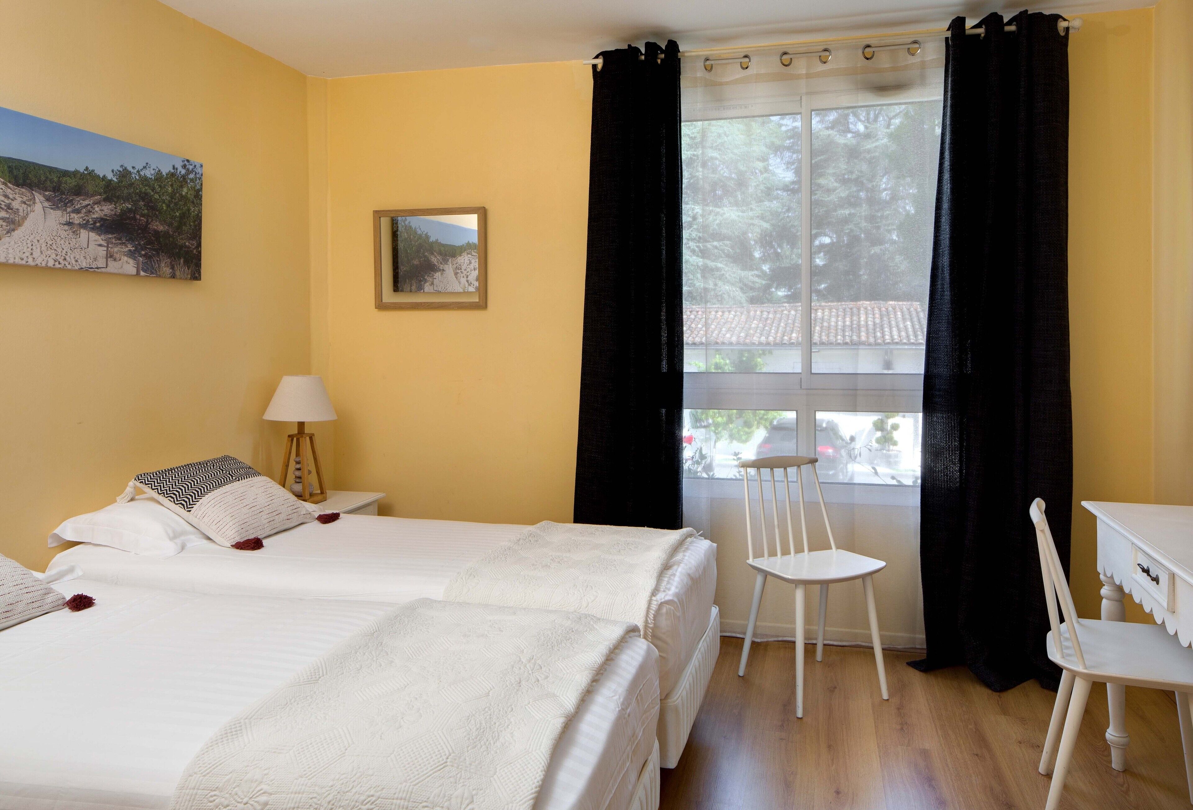 Quarto com duas camas, uma escrivaninha, uma cadeira, um abajur e uma janela com cortinas pretas.