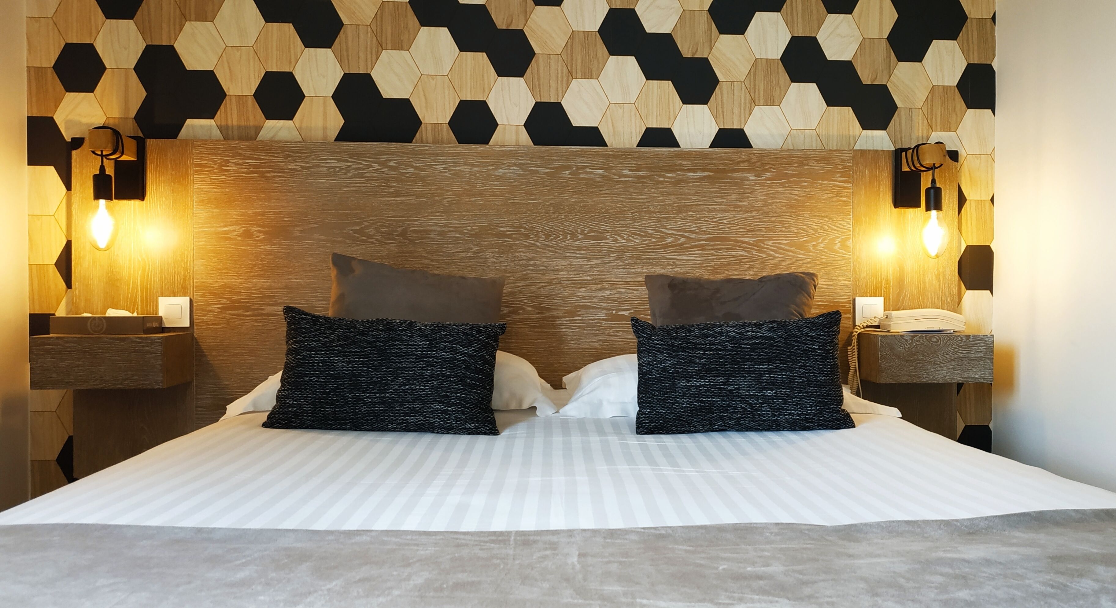 A neatly made bed with two pillows, a wooden headboard, and two wall-mounted nightstands with lamps.
