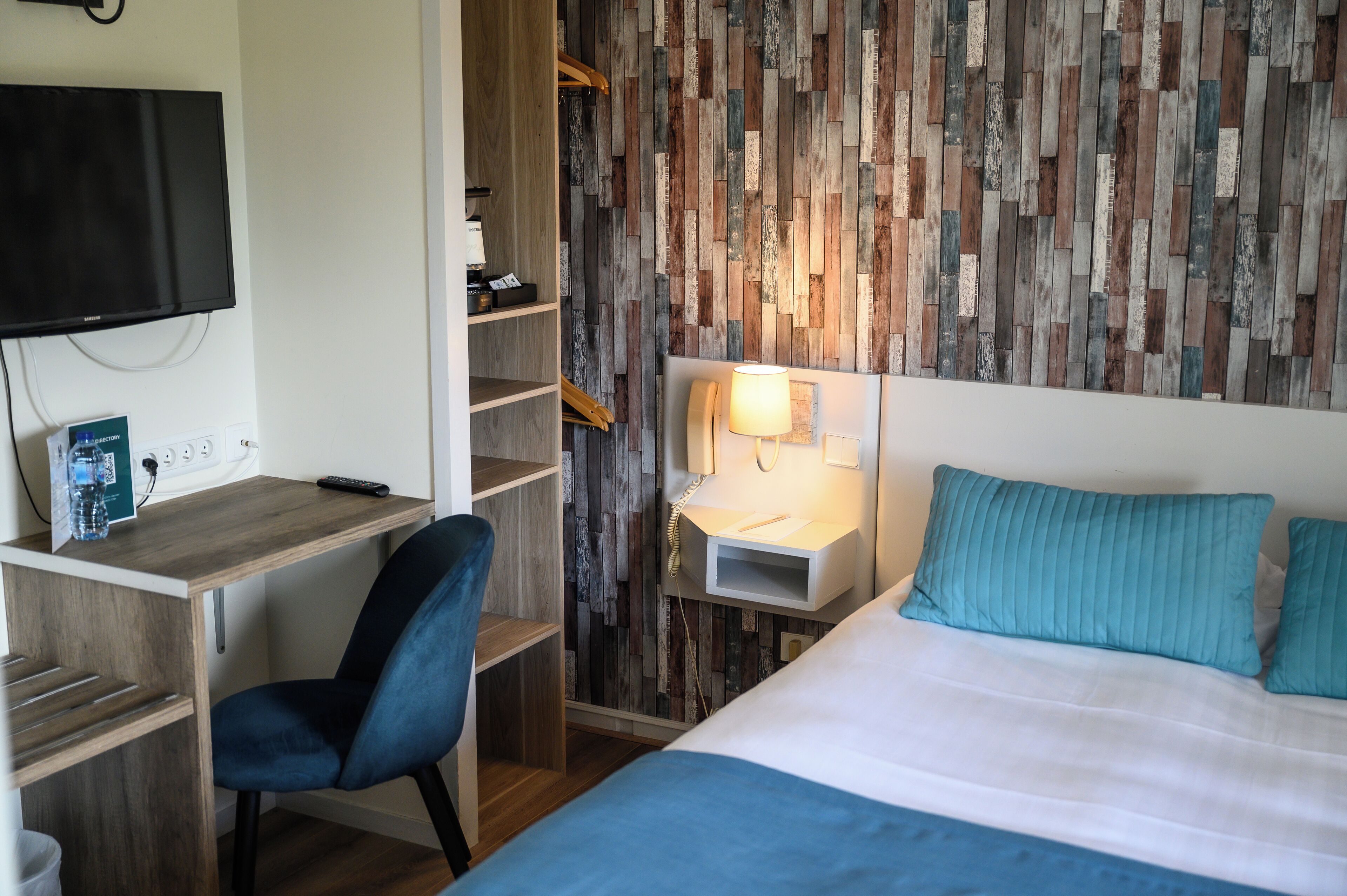 A hotel room with a wooden desk, a chair, a bed with blue bedding, a wall-mounted TV, and a wooden wall panel.