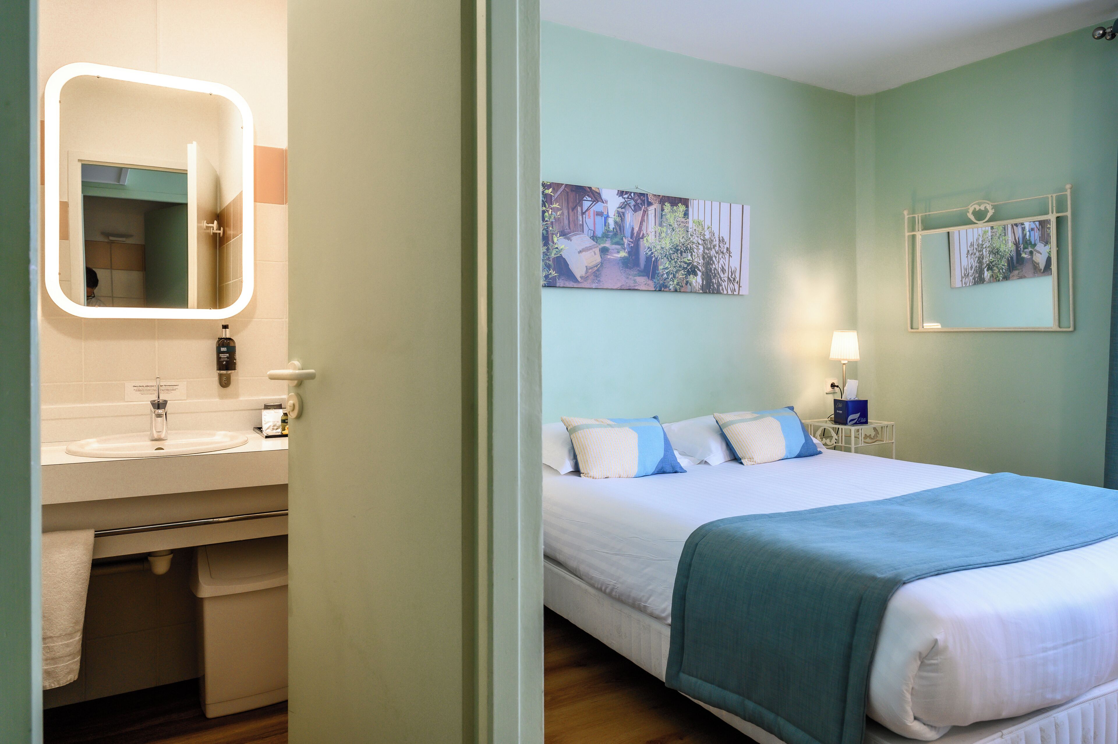 A hotel room with a bed, a bedside table with a lamp, a bathroom with a sink and mirror, and two wall-mounted pictures.