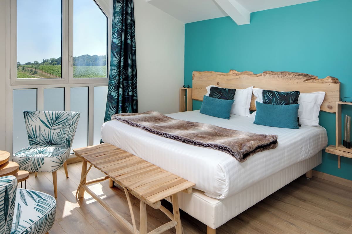 Une chambre avec un grand lit, une tête de lit en bois, un banc, une chaise et une vue sur un paysage verdoyant par la fenêtre.