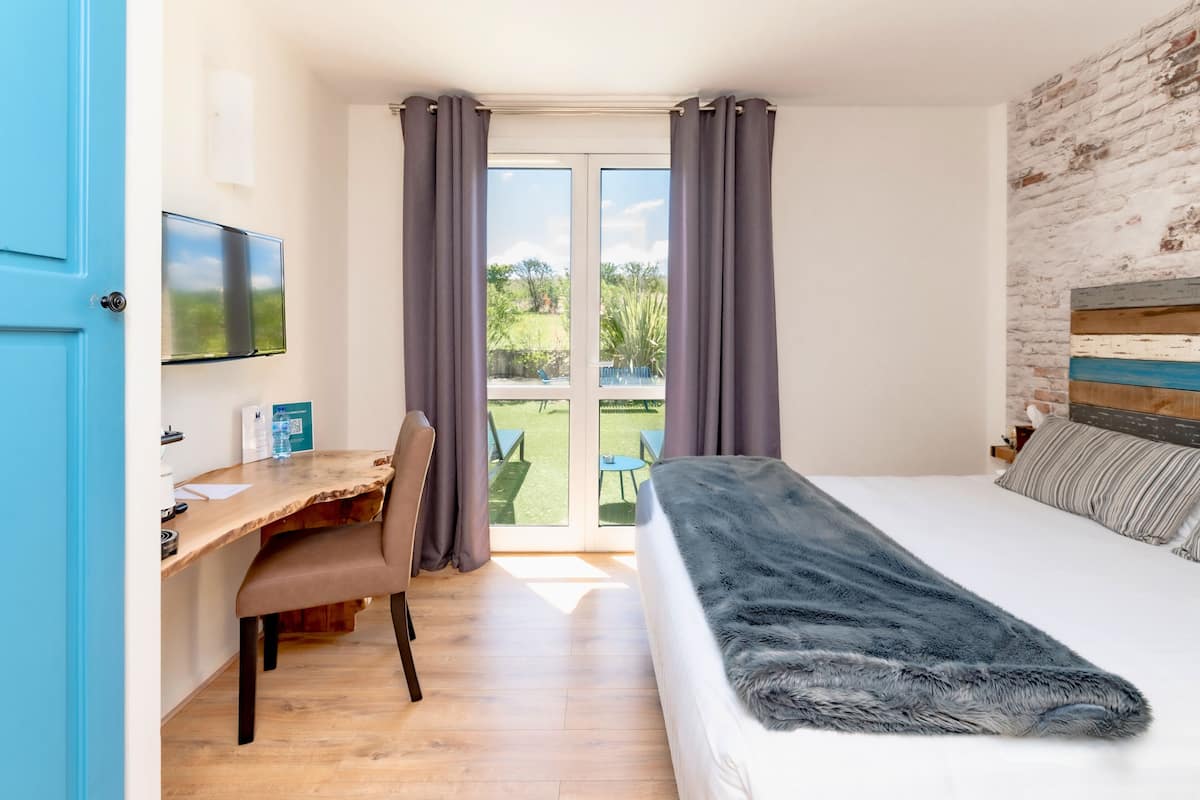 Une chambre avec un lit, un bureau et une chaise, un téléviseur fixé au mur et une vue sur l’extérieur.