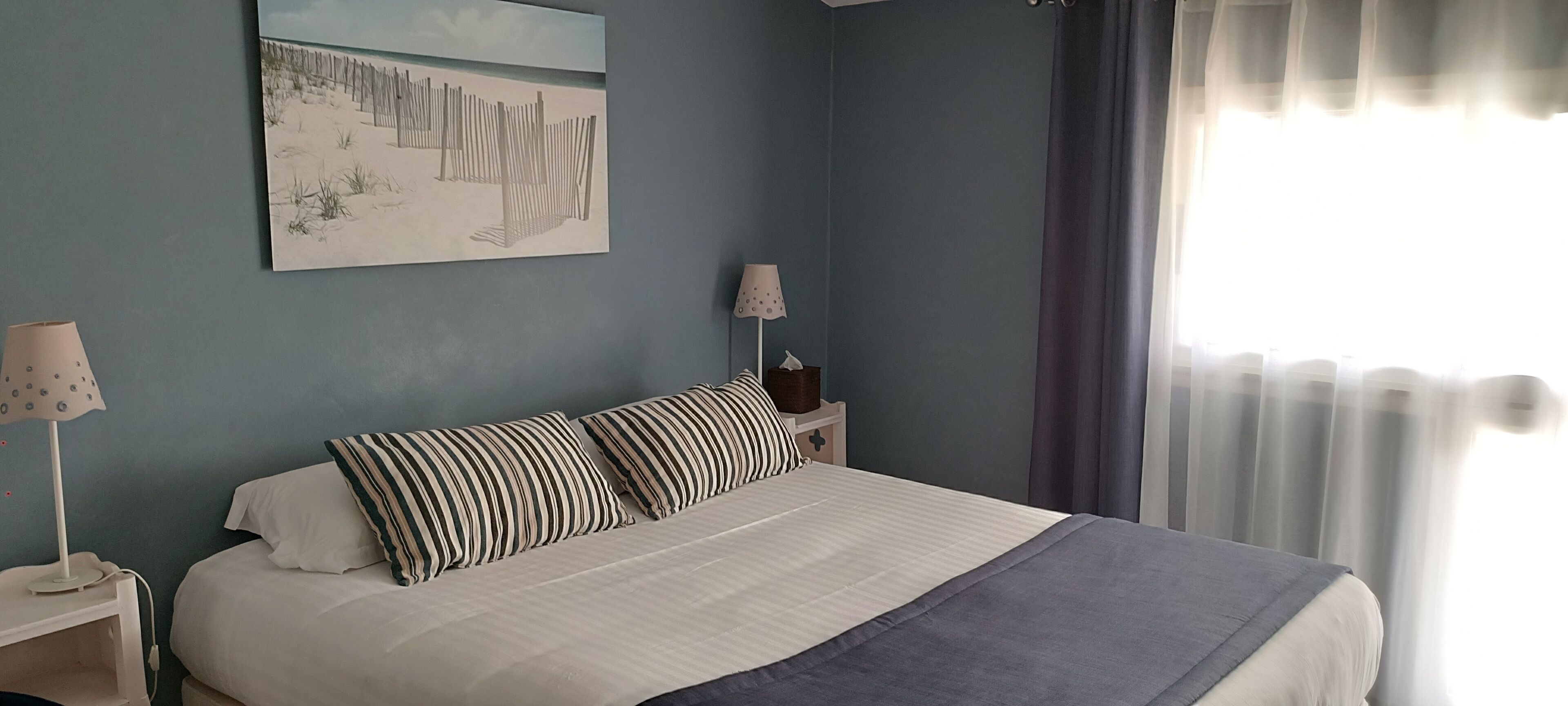 A bedroom with a bed, two bedside tables, a lamp, a painting, and a window with curtains.