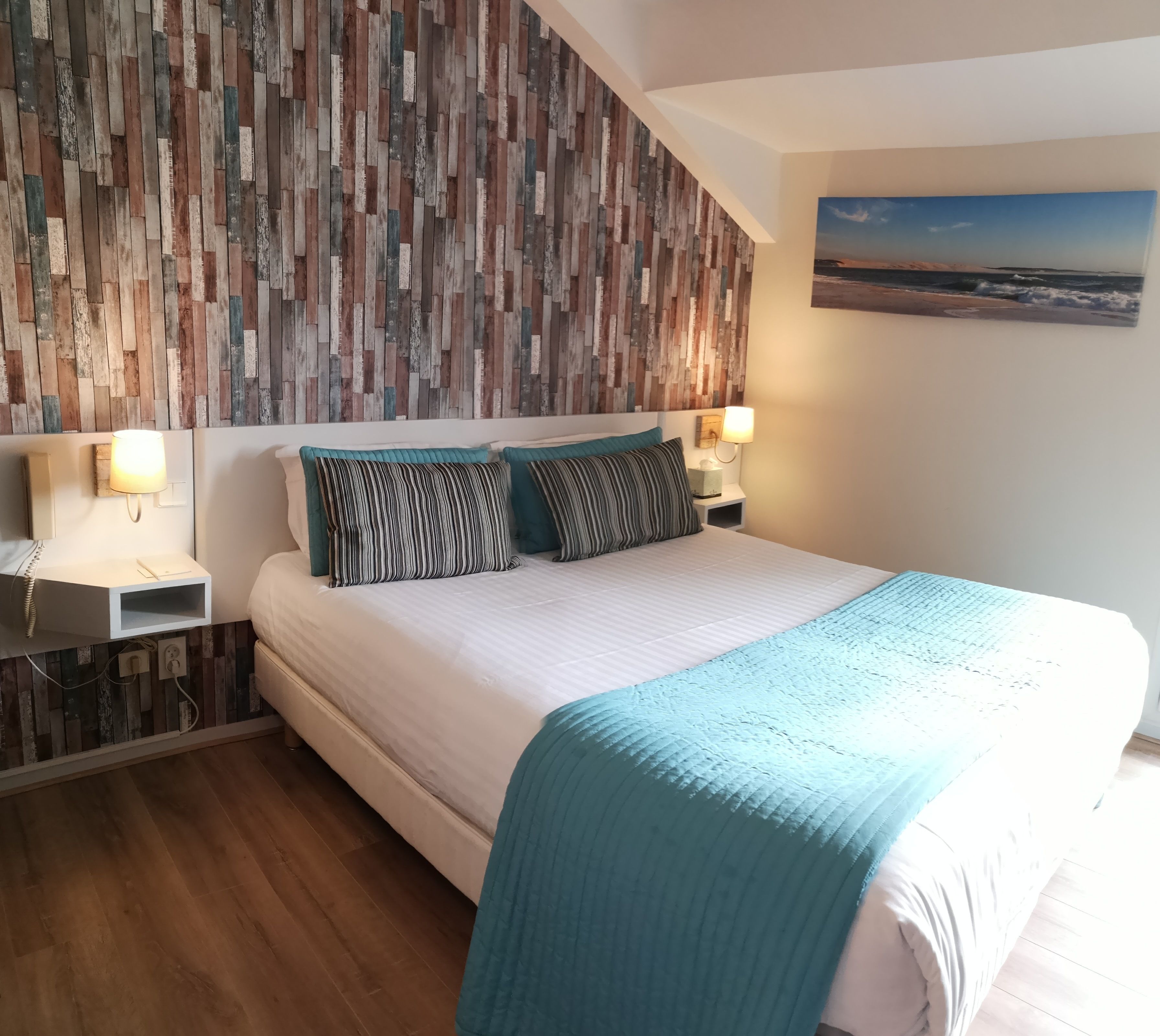 Ein Schlafzimmer mit einer Holzwandverkleidung, einem Bett mit weißer Matratze und türkisblauener Decke, zwei Nachttischen mit Lampen und einem Gemälde an der Wand.