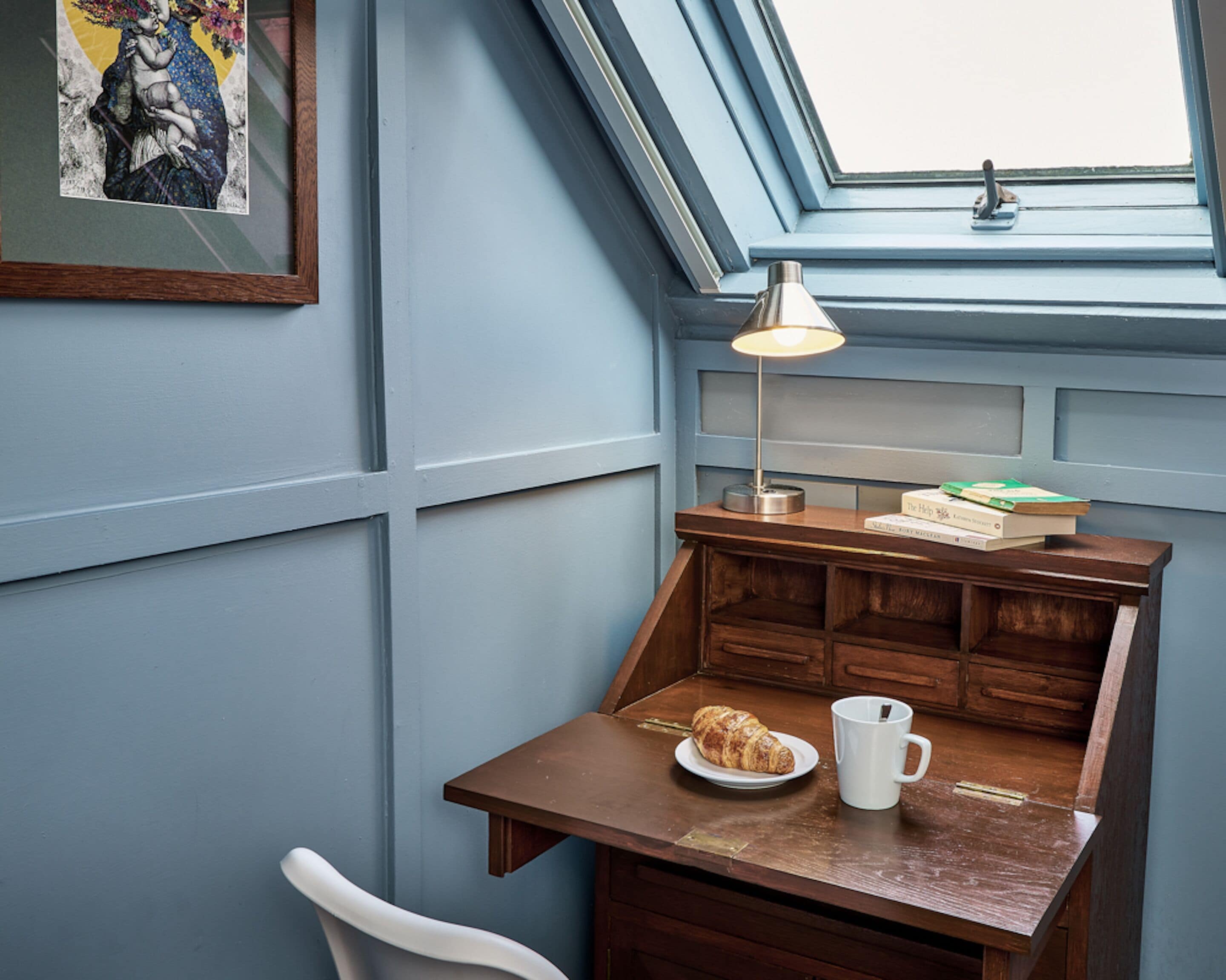 Un coin douillet avec un bureau en bois, une lampe, une assiette contenant un croissant, une tasse et un tableau encadré accroché au mur.