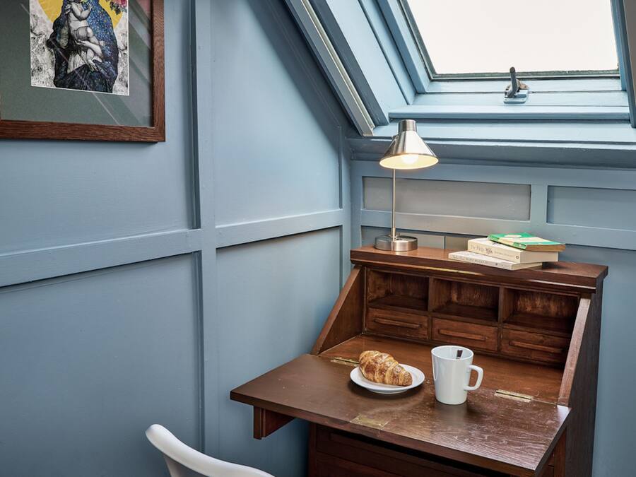 A cozy corner with a wooden desk, a lamp, a plate with a croissant, a mug, and a framed picture on the wall.