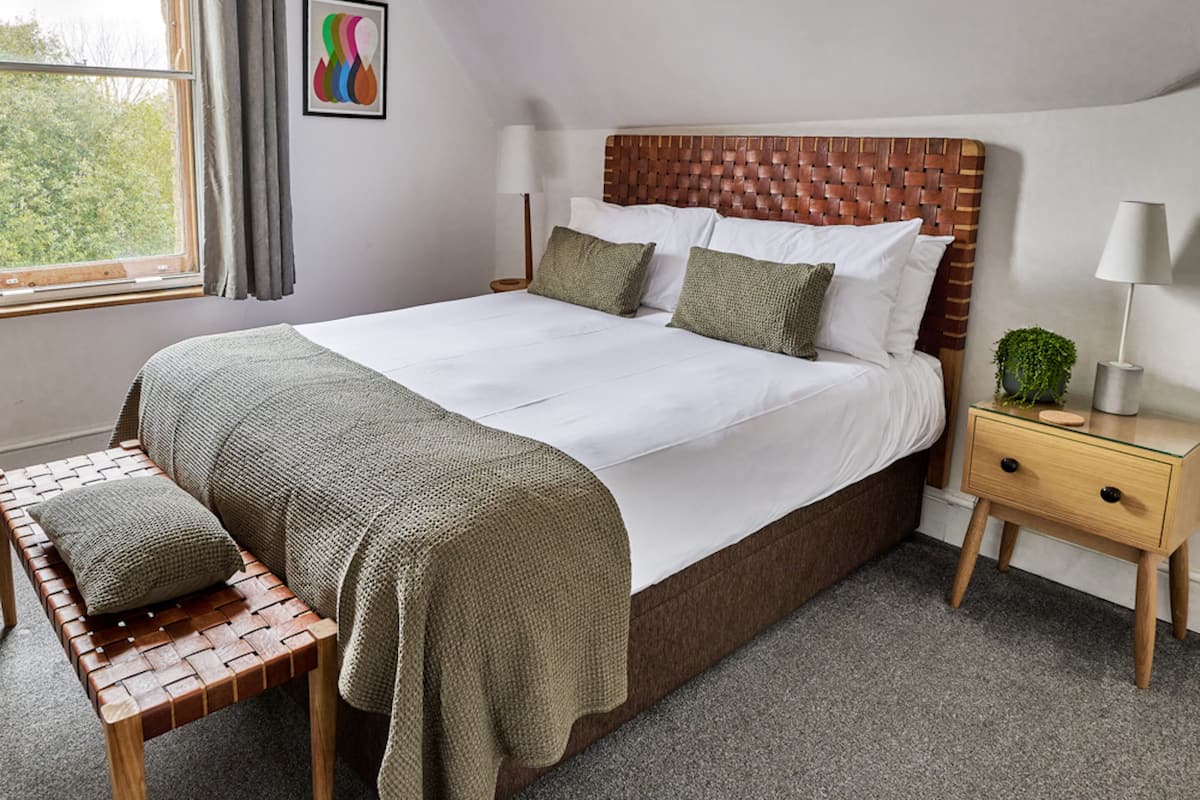 A neatly made bed with a woven headboard, a bench with a woven cover, a bedside table with a lamp, and a framed picture on the wall.