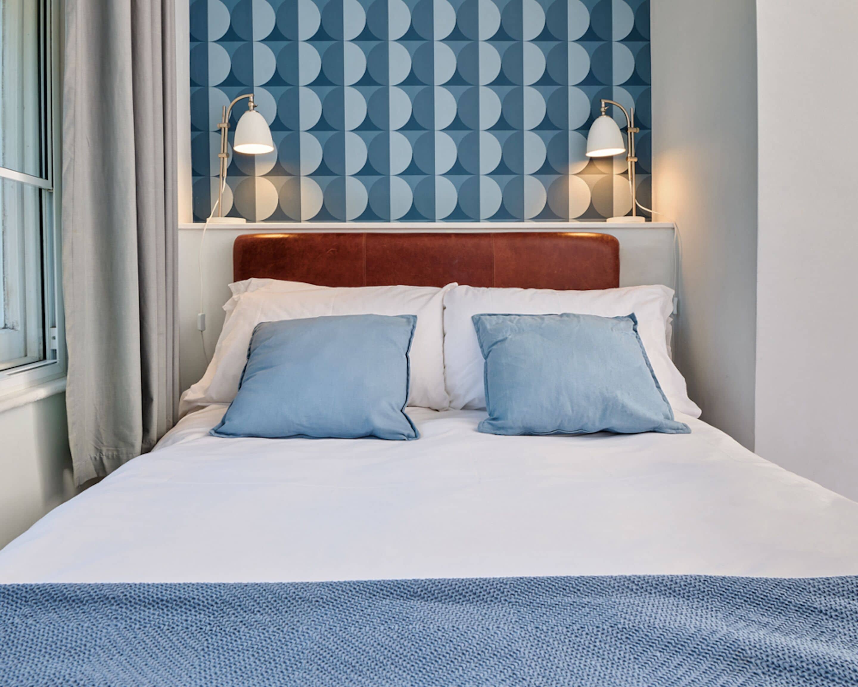 A neatly made bed with two pillows, a headboard, and two wall-mounted lamps.