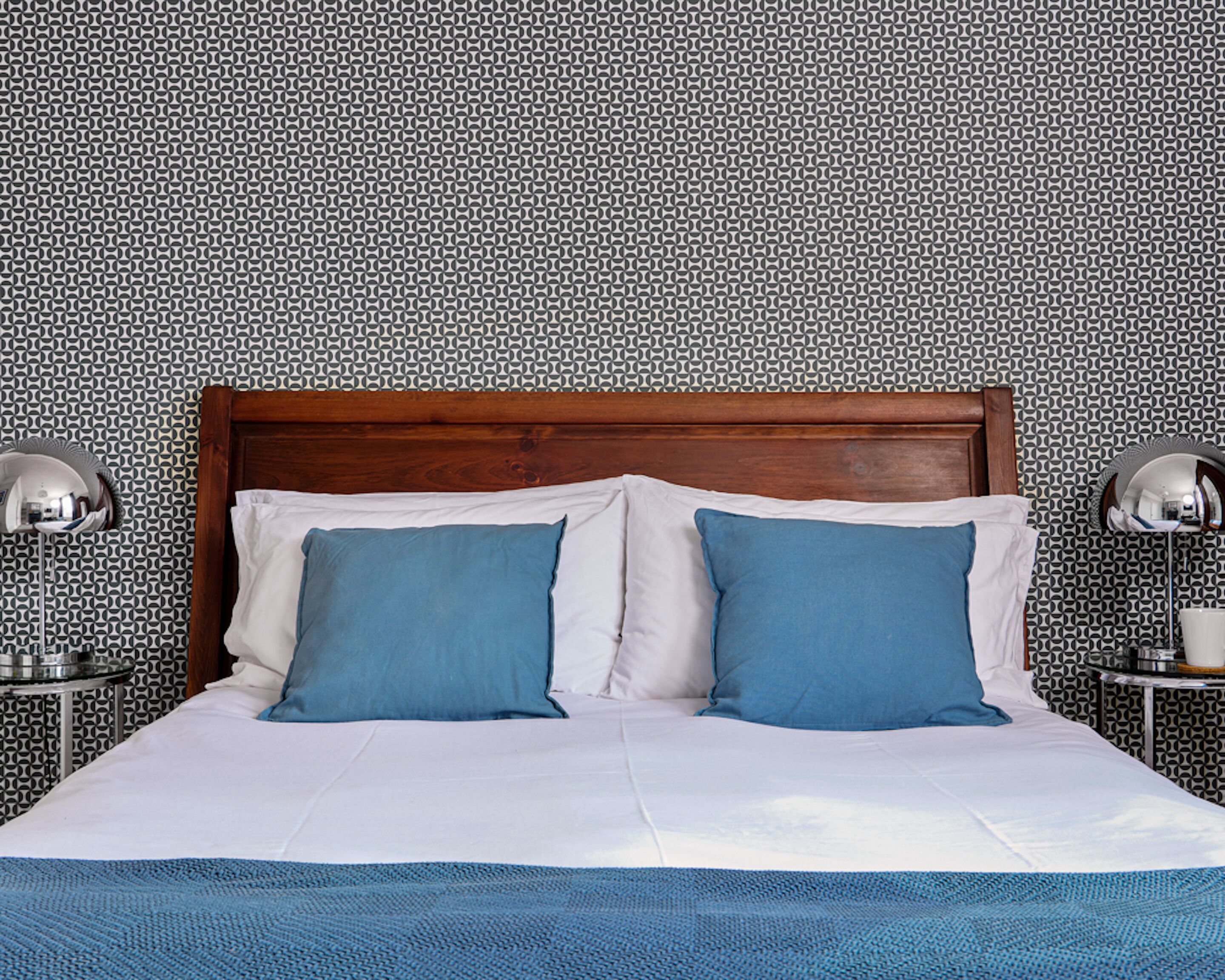 Una cama bien hecha con cabecera de madera, dos almohadas y una colcha azul. Hay dos mesitas de noche con lámparas a cada lado de la cama.
