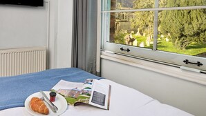 Habitación de hotel con cama, ventana con vista a un cementerio y árboles, televisor de pantalla plana en la pared, y una mesa con un croissant, café y una revista.