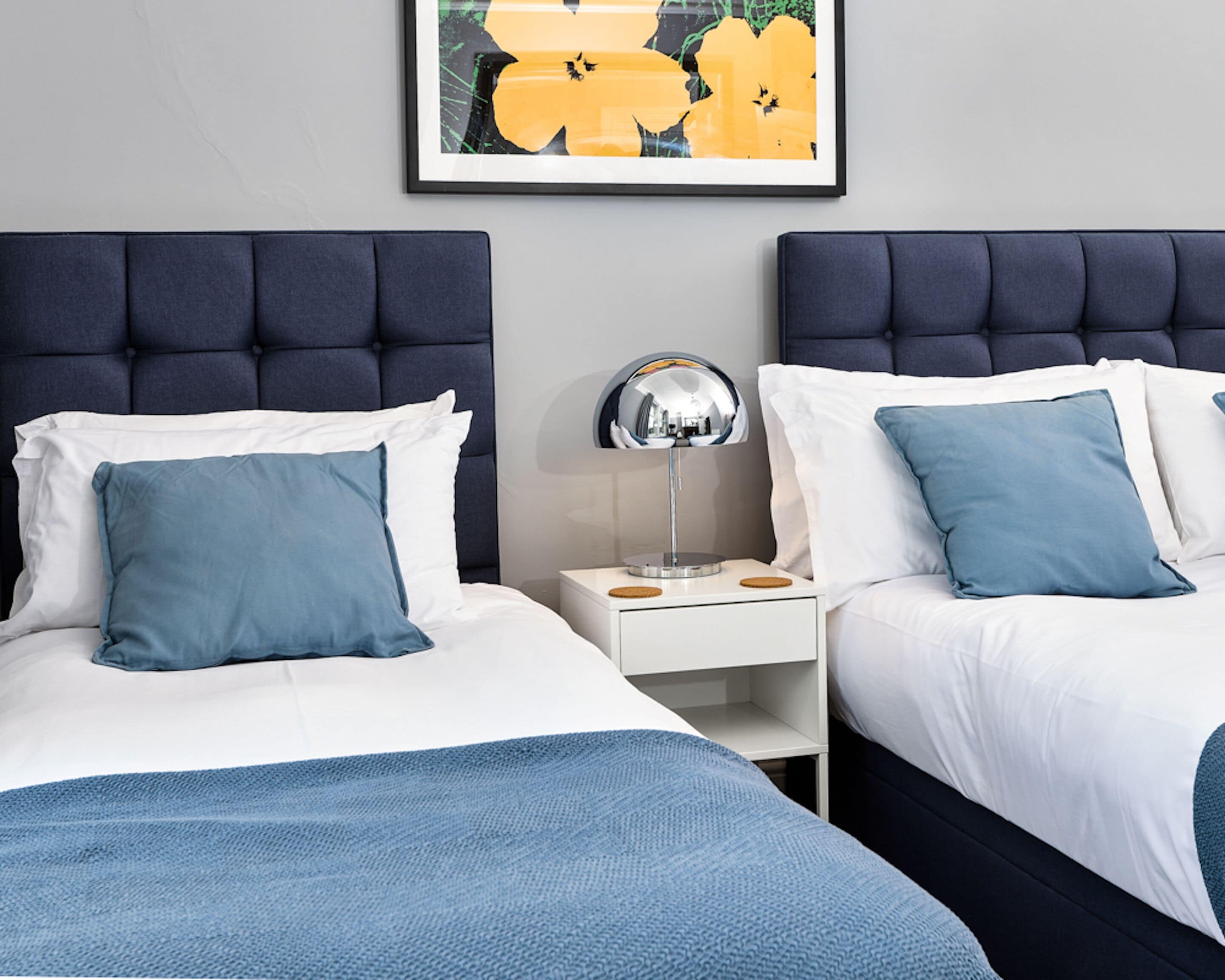 A hotel room with two beds, each with a dark headboard, white bedding, and blue pillows. There is a nightstand with a mirror and a small table between the beds. A framed artwork is visible on the wall above the beds.