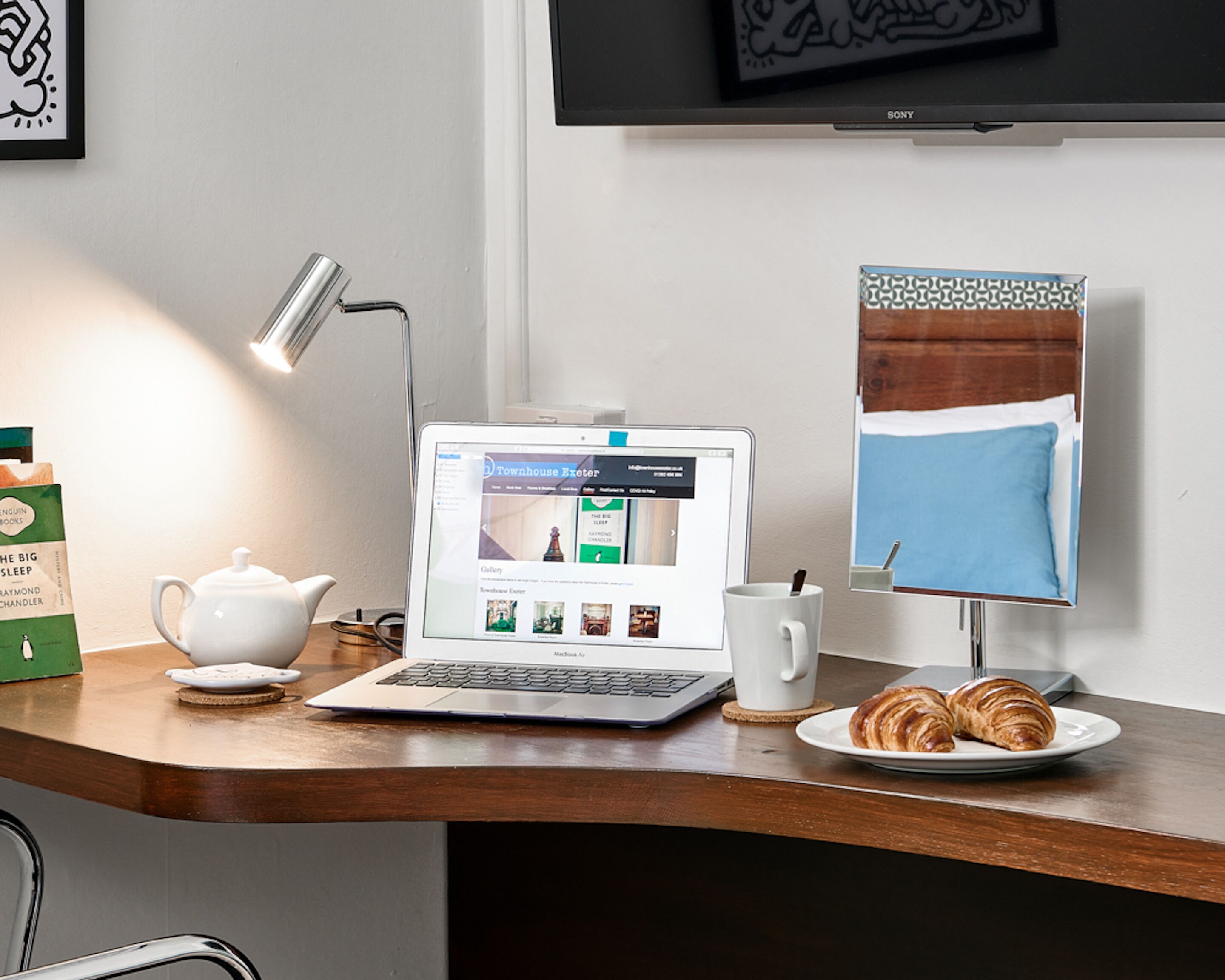 Una laptop sobre un escritorio de madera con una taza, una tetera y un plato con cruasanes.