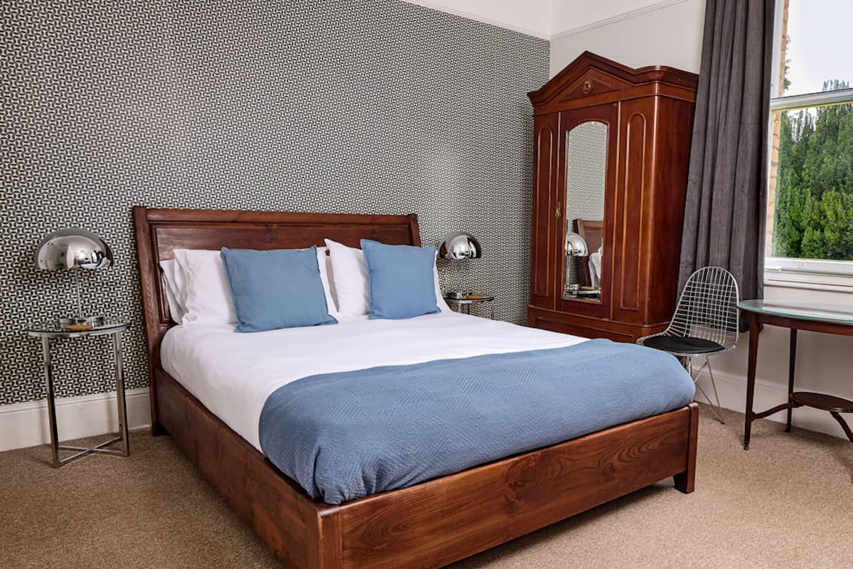 A neatly made bed with a wooden frame, two bedside tables with lamps, a large mirror, and a view of greenery through a window.