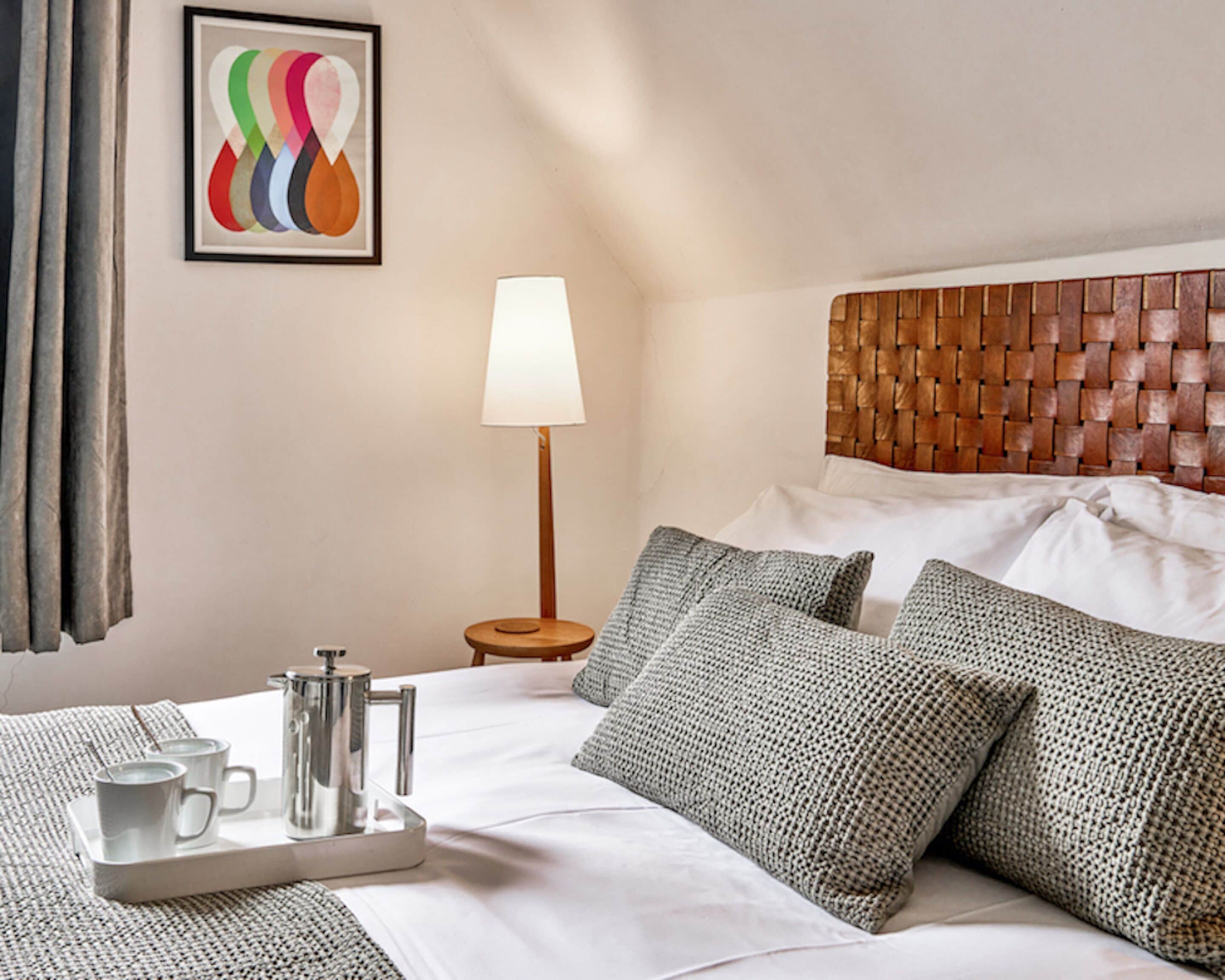 Une chambre d’hôtel avec un lit soigneusement fait, une table de chevet avec une lampe, un plateau avec des tasses et une carafe, et une peinture abstraite aux couleurs vives accrochée au mur.