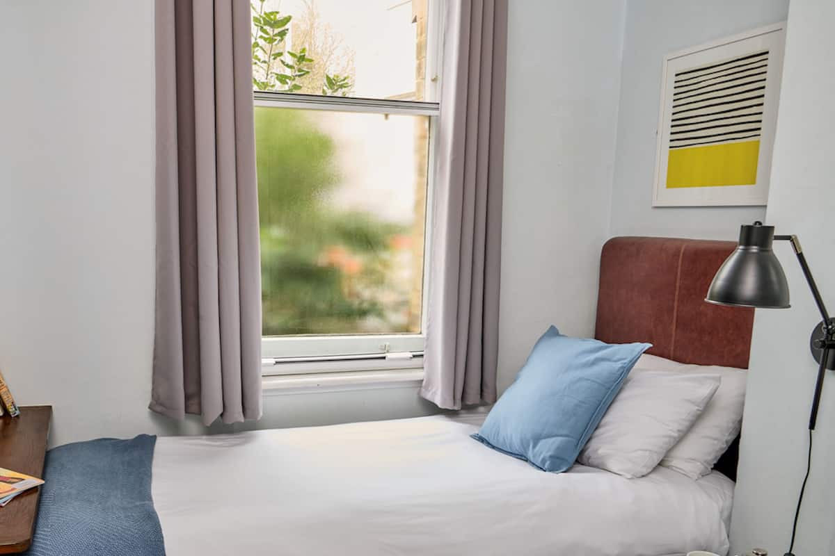 A neatly made bed with white and blue bedding, a bedside table with a lamp, and a window with curtains.