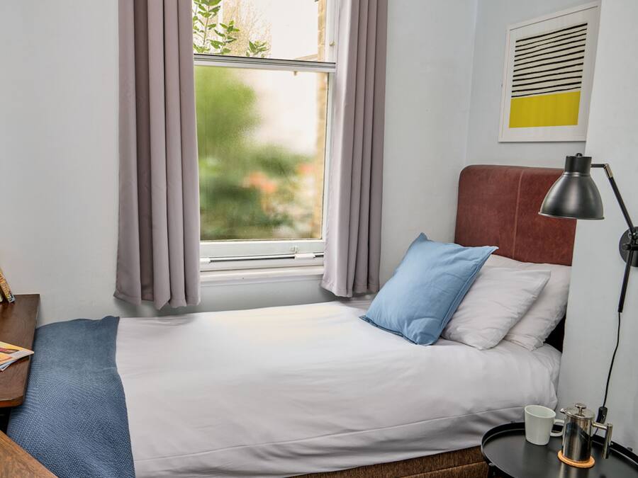 A neatly made bed with white and blue bedding, a bedside table with a lamp, and a window with curtains.