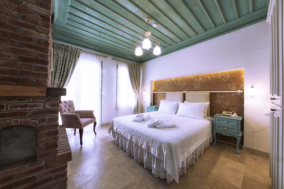 A bedroom with a brick fireplace, a large bed with white linens, a nightstand, and a chair.